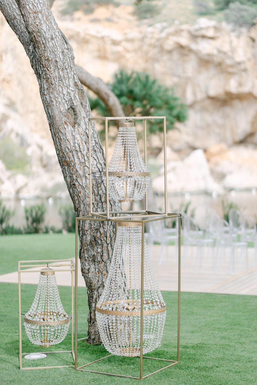 Crystal chandeliers hanging from metal stands near a tree trunk in an outdoor garden setting.