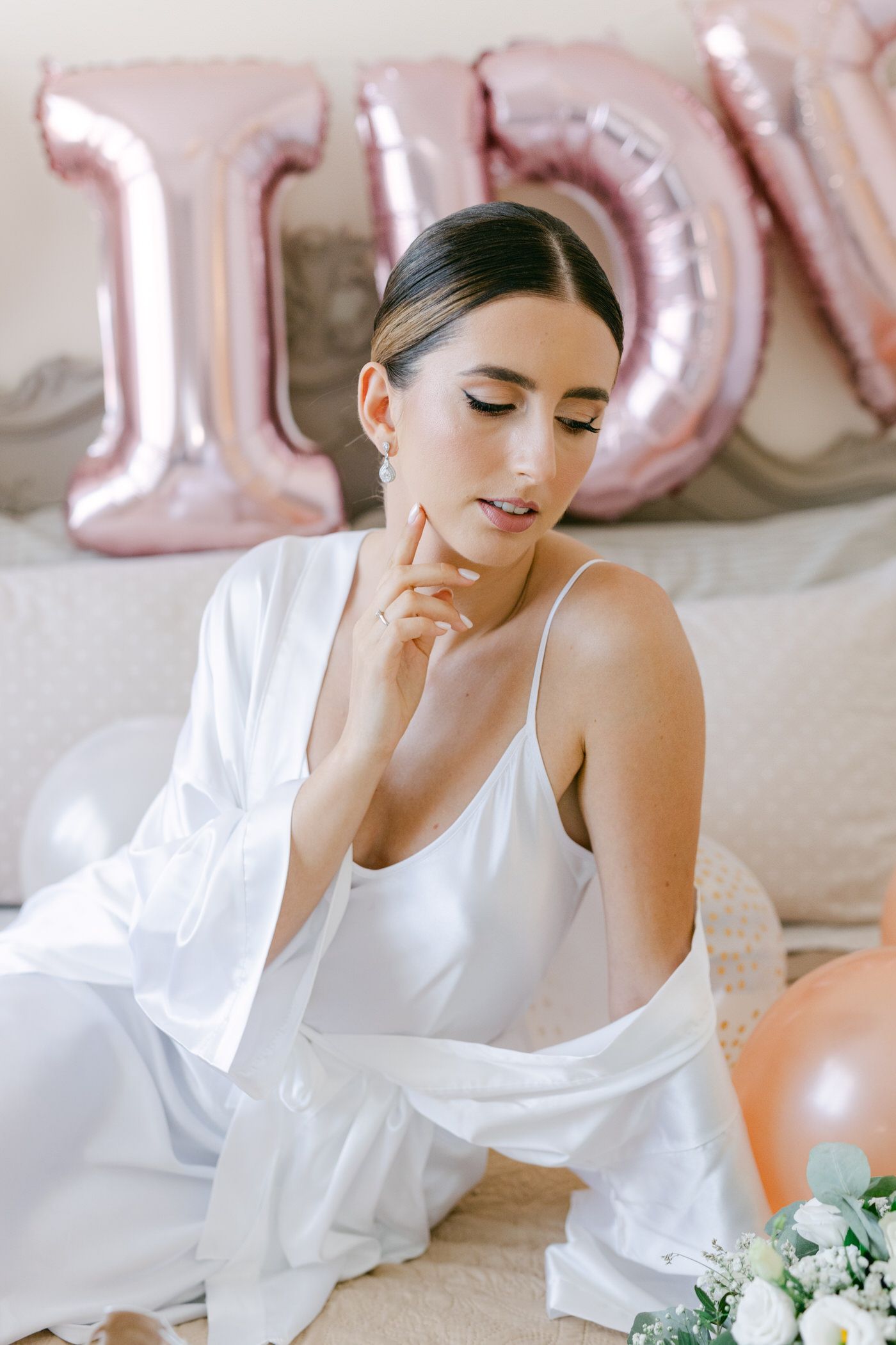 Woman in white sleepwear sitting thoughtfully against pink balloon decorations and floral accents.