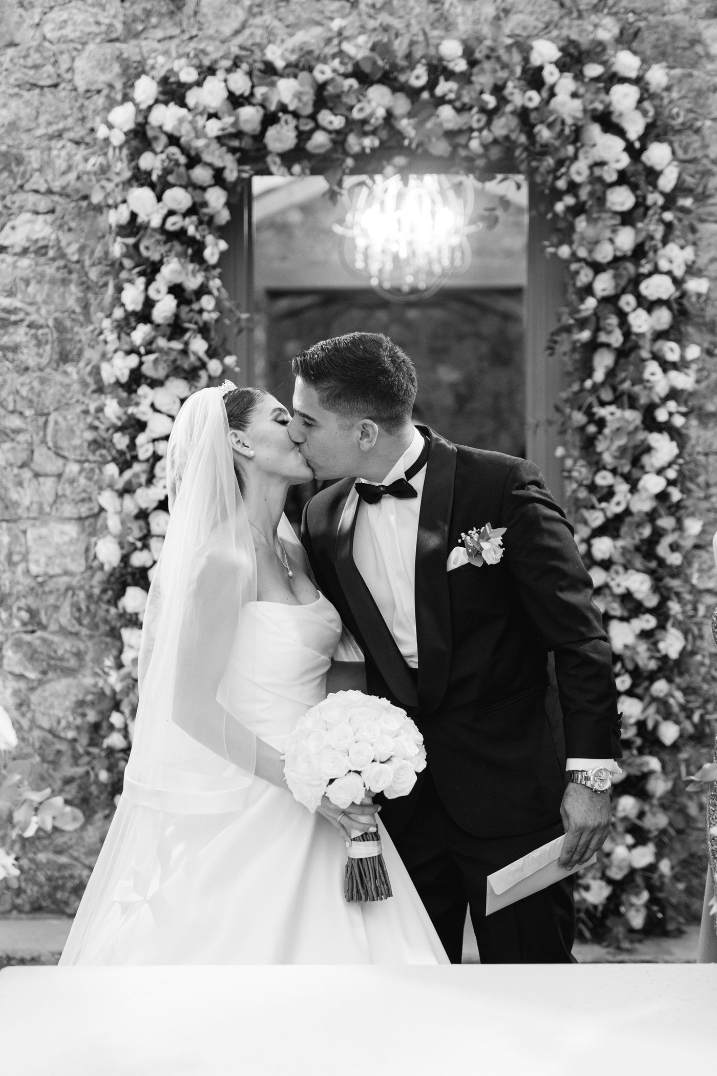 Romantic black and white wedding photo under a floral archway with stone wall background.