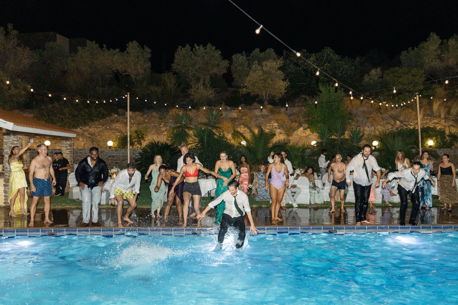 Wedding guests celebrate around a pool with string lights overhead at night.