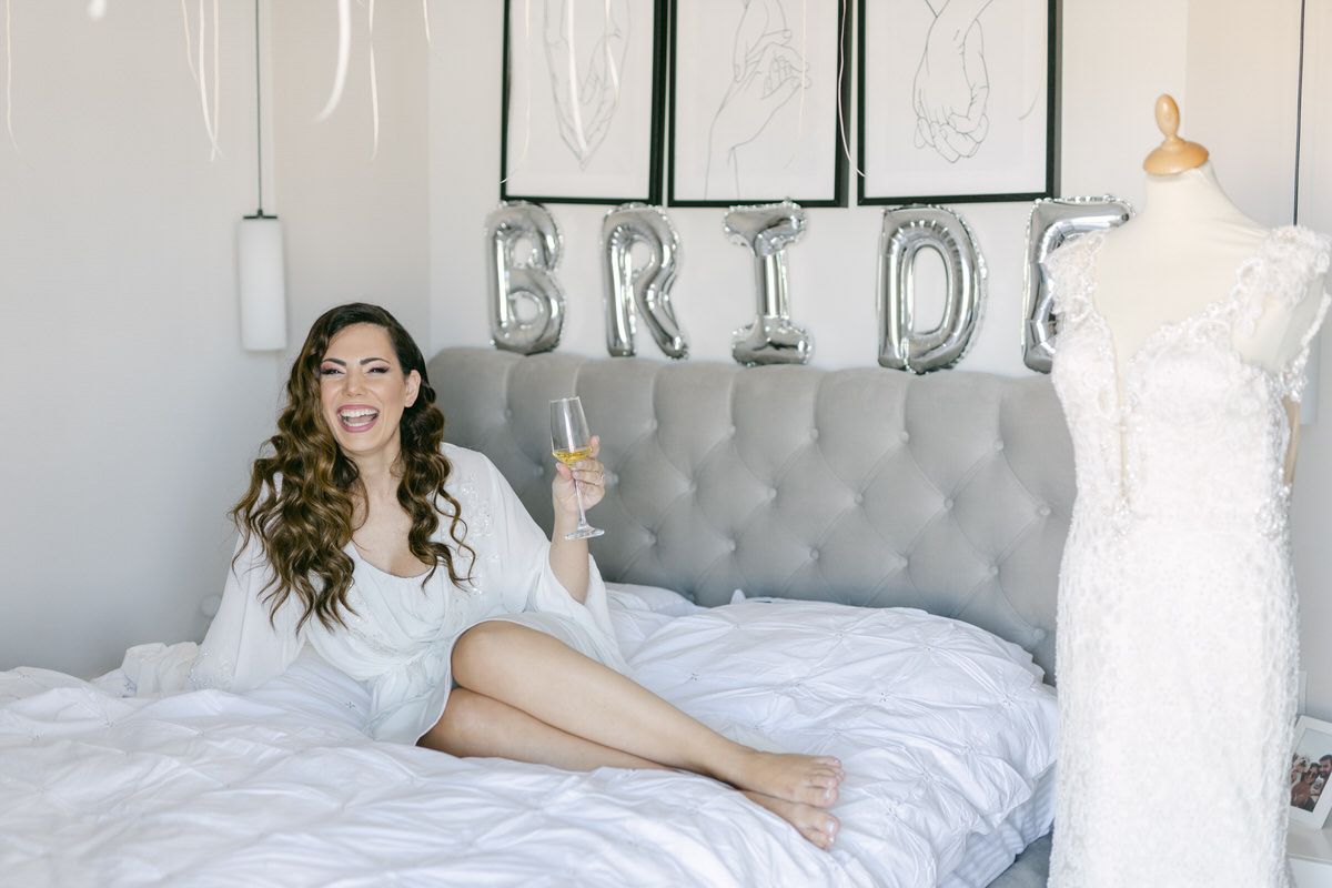 A bridal preparation scene with silver BRIDE letter balloons, a gray tufted headboard, and a wedding dress on display.