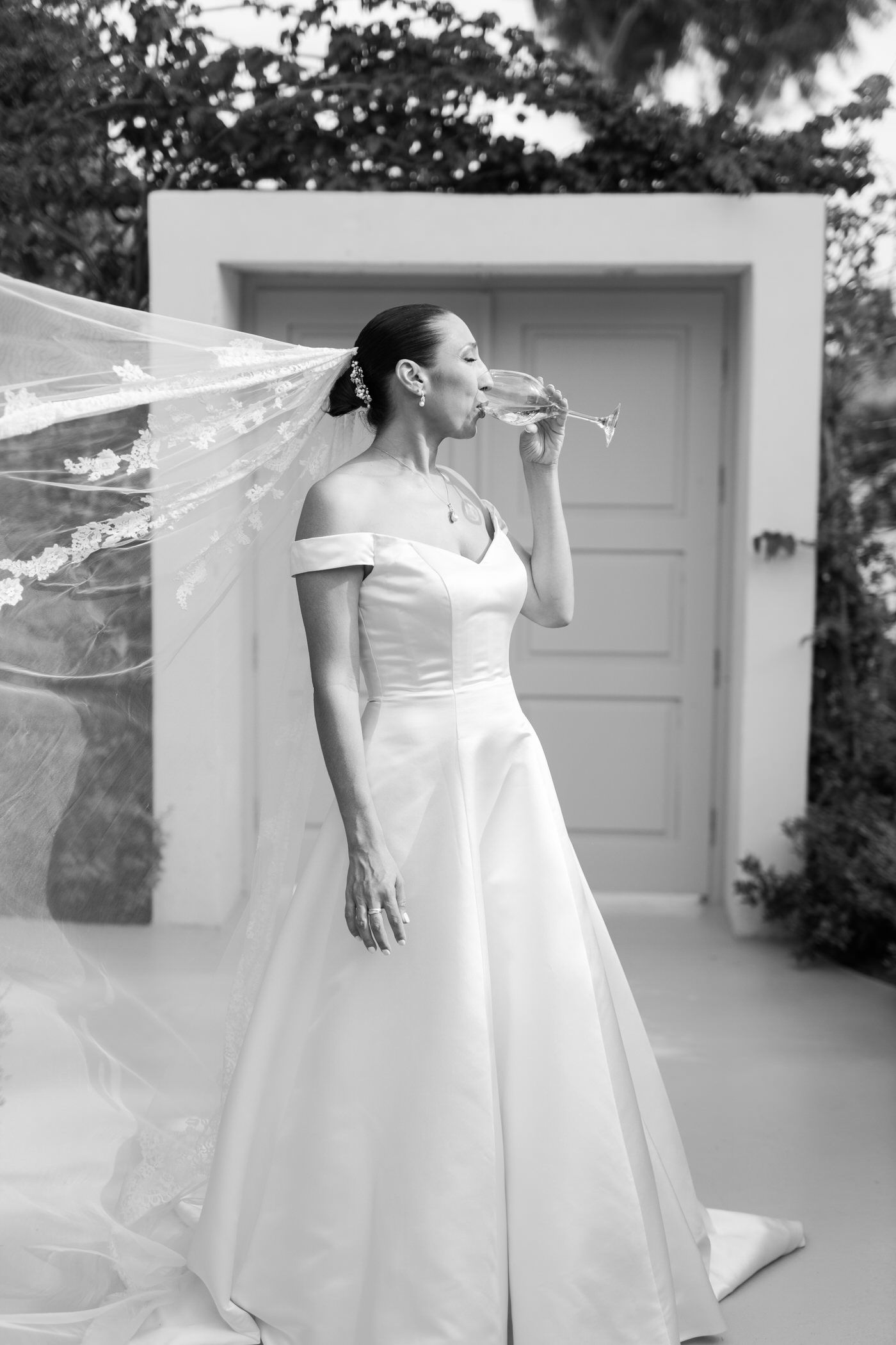 Elegant bride in off-shoulder white gown and flowing veil stands gracefully in front of white door in black and white photo.