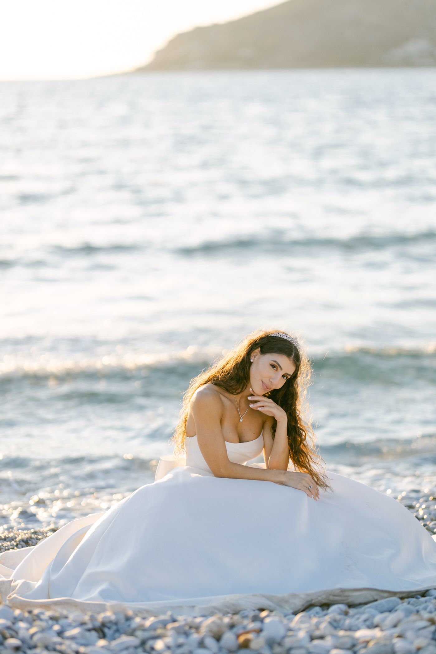 A woman in a white wedding gown sits on a rocky beach at sunset with ocean waves in the background.