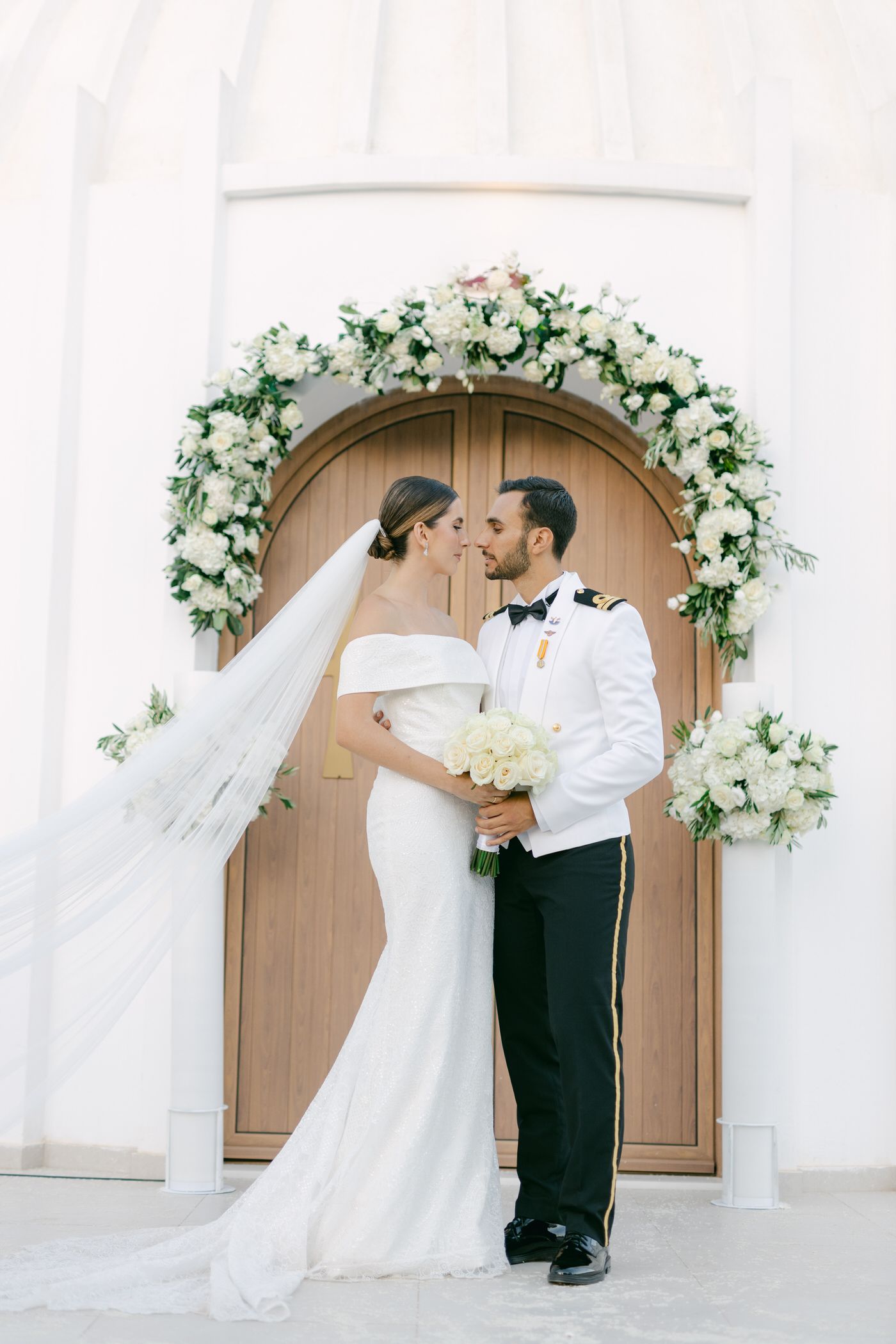 A romantic wedding couple stands beneath an elegant white floral arch against a wooden church door.