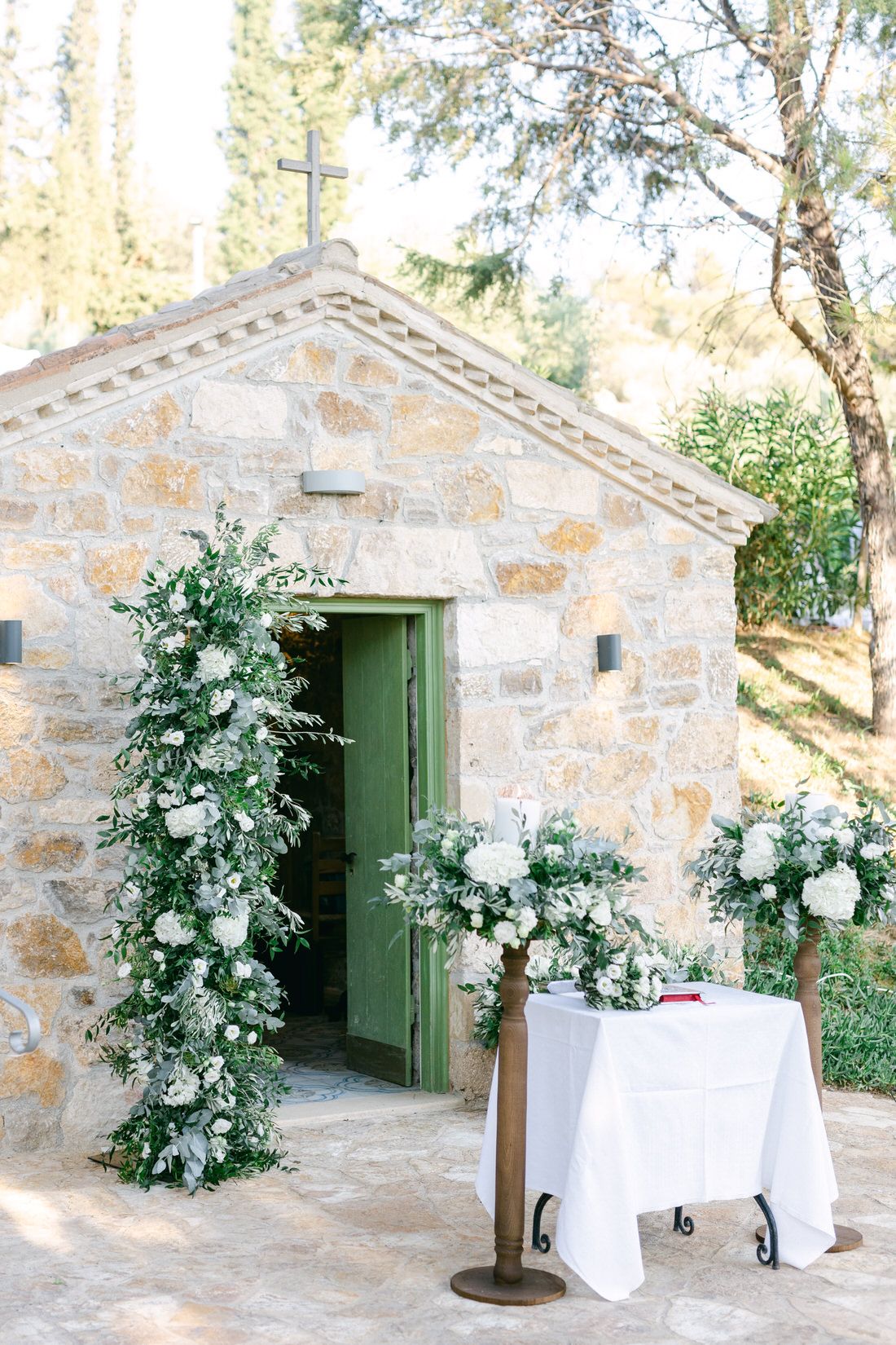 Rustic stone building entrance decorated with elegant white floral arrangements and green foliage around a green wooden door.
