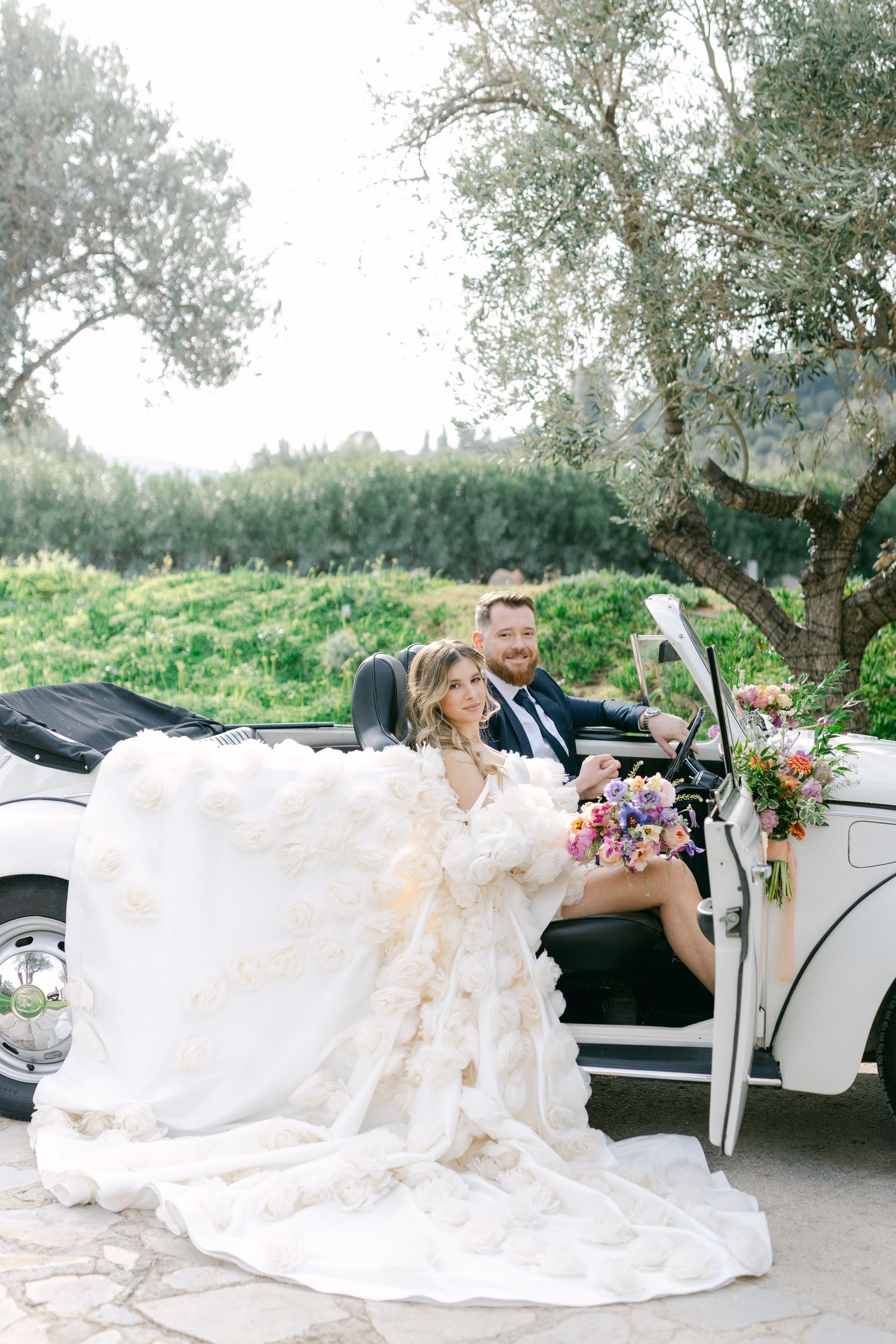 A couple sits in a vintage white convertible car with a flowing wedding dress and pink floral bouquet in a garden setting.