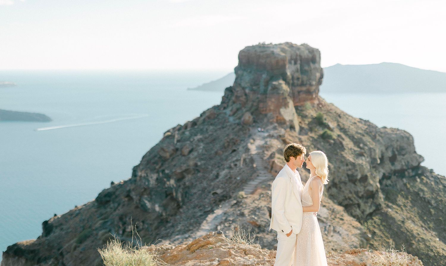A couple in white embraces on a scenic clifftop overlooking the Mediterranean waters of Santorini, Greece.