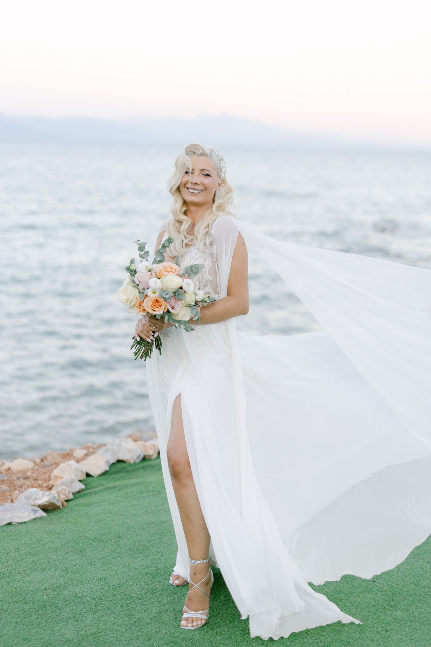 A bride in a flowing white wedding dress with a thigh-high slit holds a peach and green bouquet by the ocean.