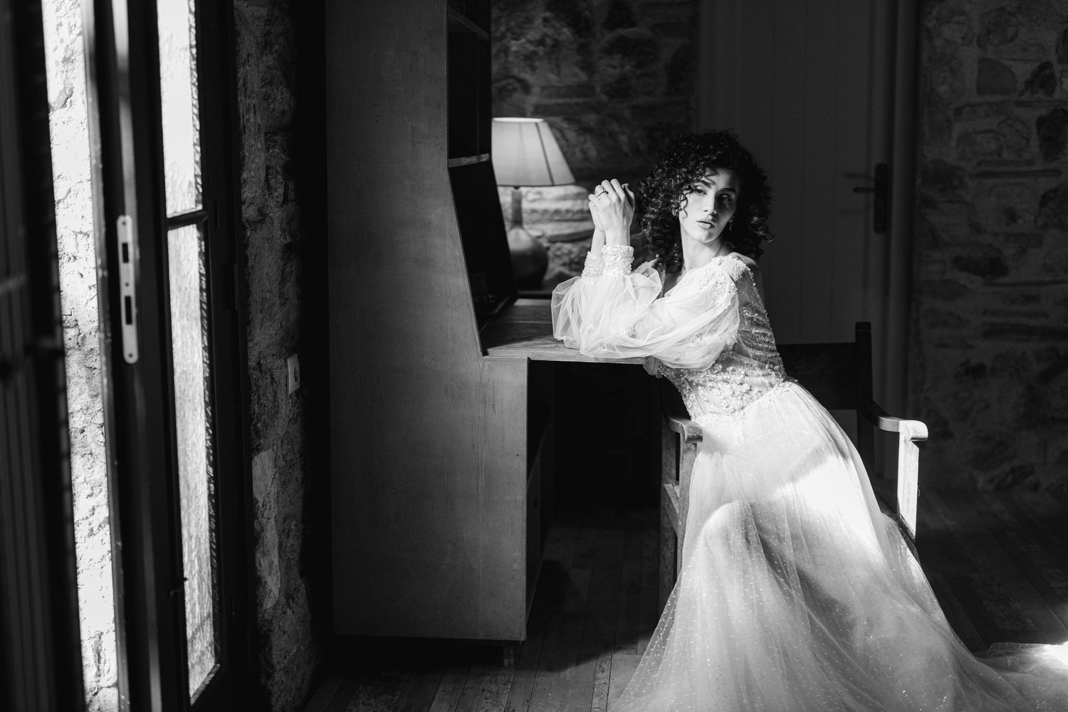 A bride in a flowing white wedding dress poses dramatically by a window in black and white photography.