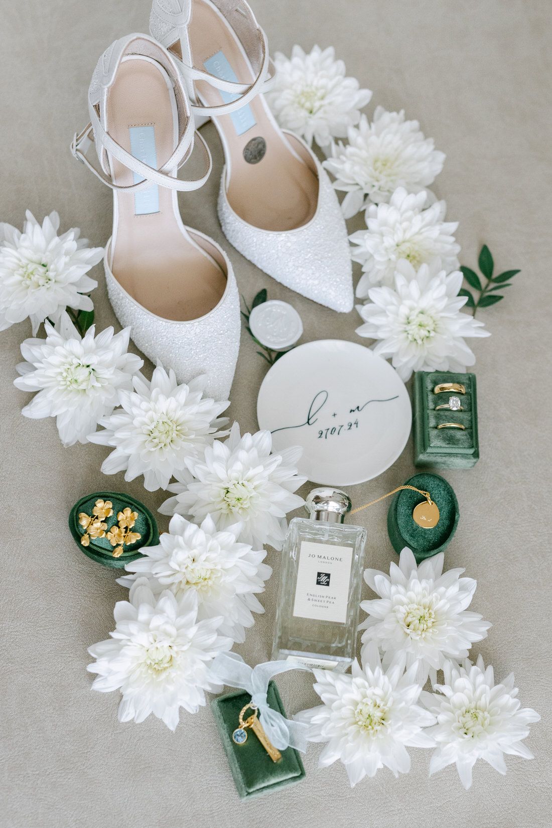 Bridal accessories flatlay with white heels, flowers, perfume bottles and jewelry on beige background.