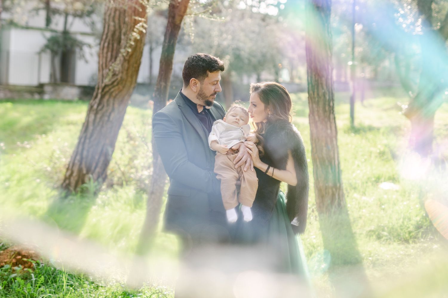 A couple shares a tender moment with their baby in a sunlit park with trees while rainbow light flares dance across the frame.