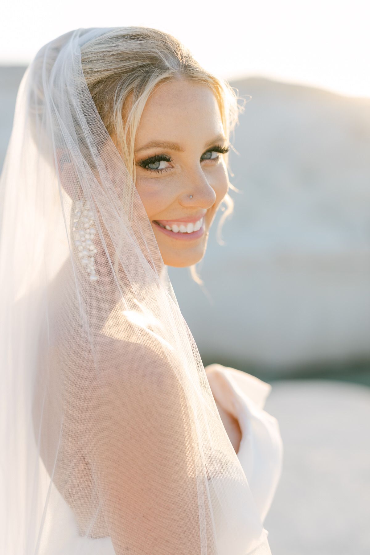 A bride smiles radiantly in golden sunlight wearing a white veil and strapless wedding dress.