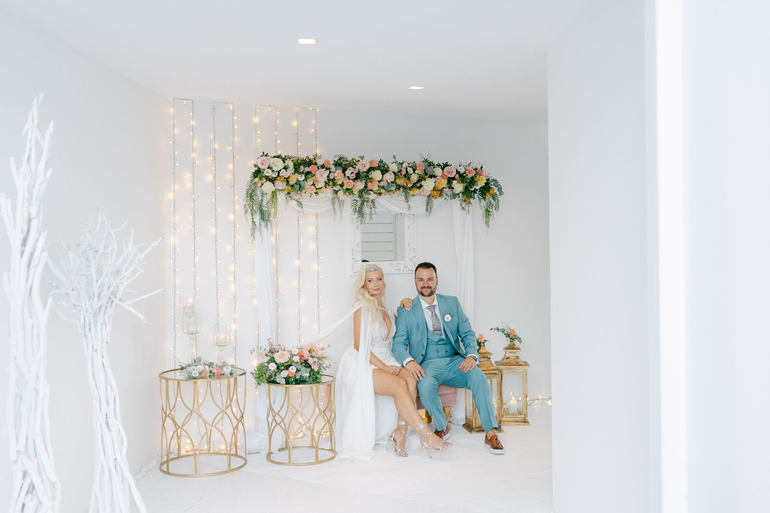 A couple sits in an elegant white room with floral decor, fairy lights, and golden geometric stands for a wedding photoshoot.