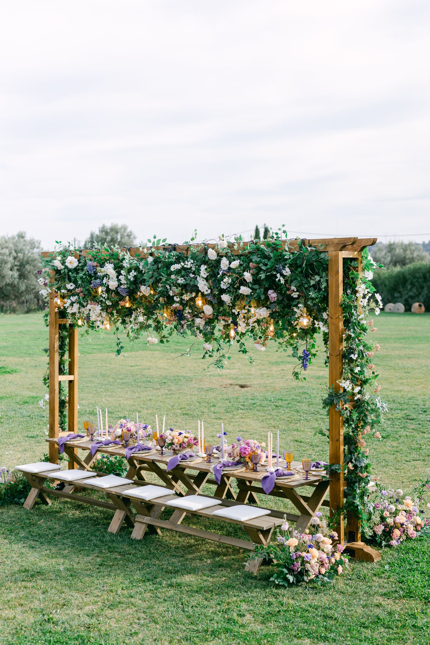 Rustic wooden picnic table setup under floral arch with pink and purple table decor in outdoor garden setting.