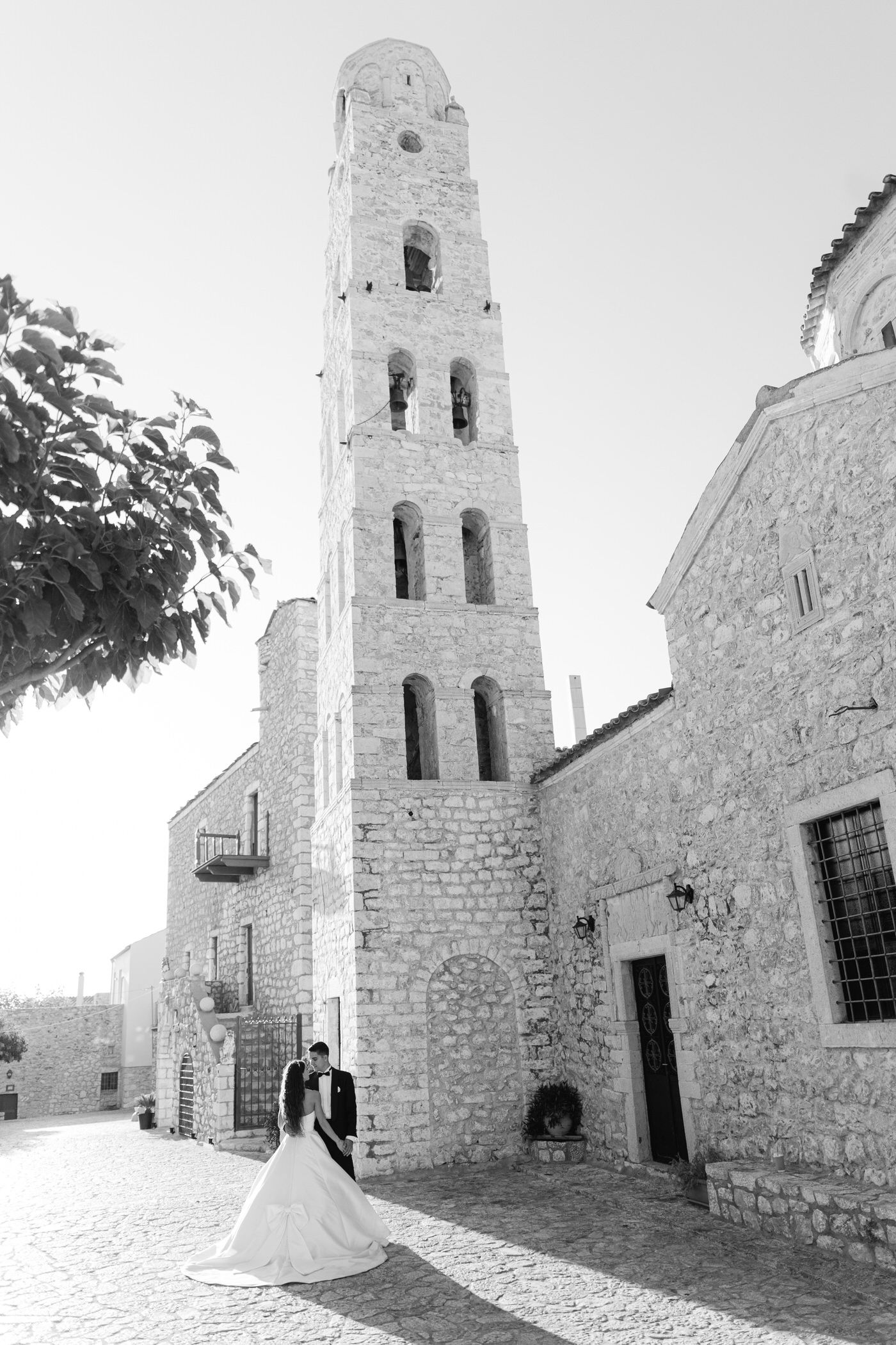 A romantic wedding photo at a historic stone church with a tall bell tower and cobblestone courtyard in black and white.