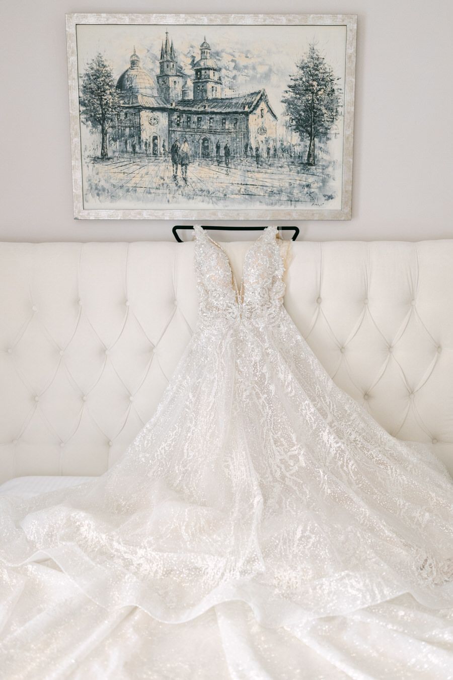 Delicate lace wedding gown displayed on white tufted sofa beneath vintage artwork.