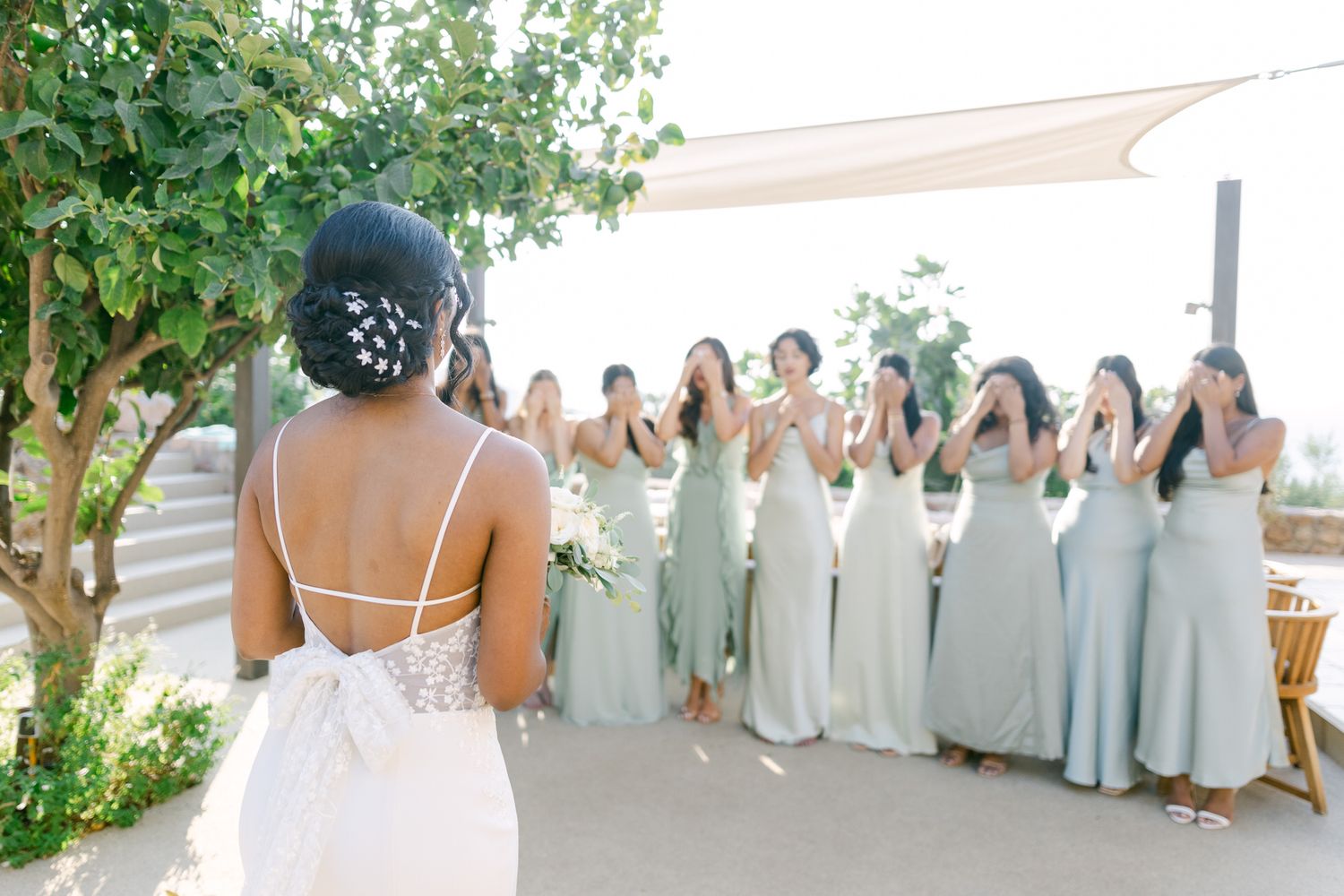 Bridesmaids in pale sage green dresses gather together behind a bride in a white wedding gown.