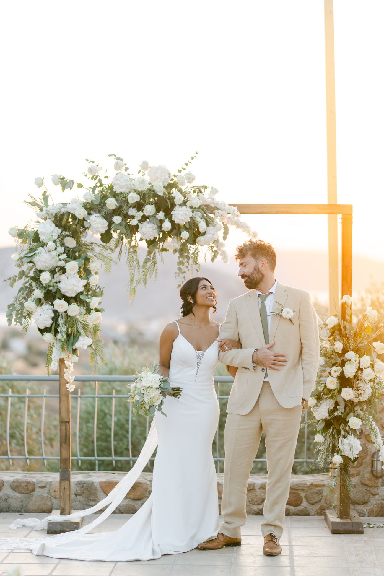 Newlyweds stand under a floral arch with white roses and greenery during golden hour.