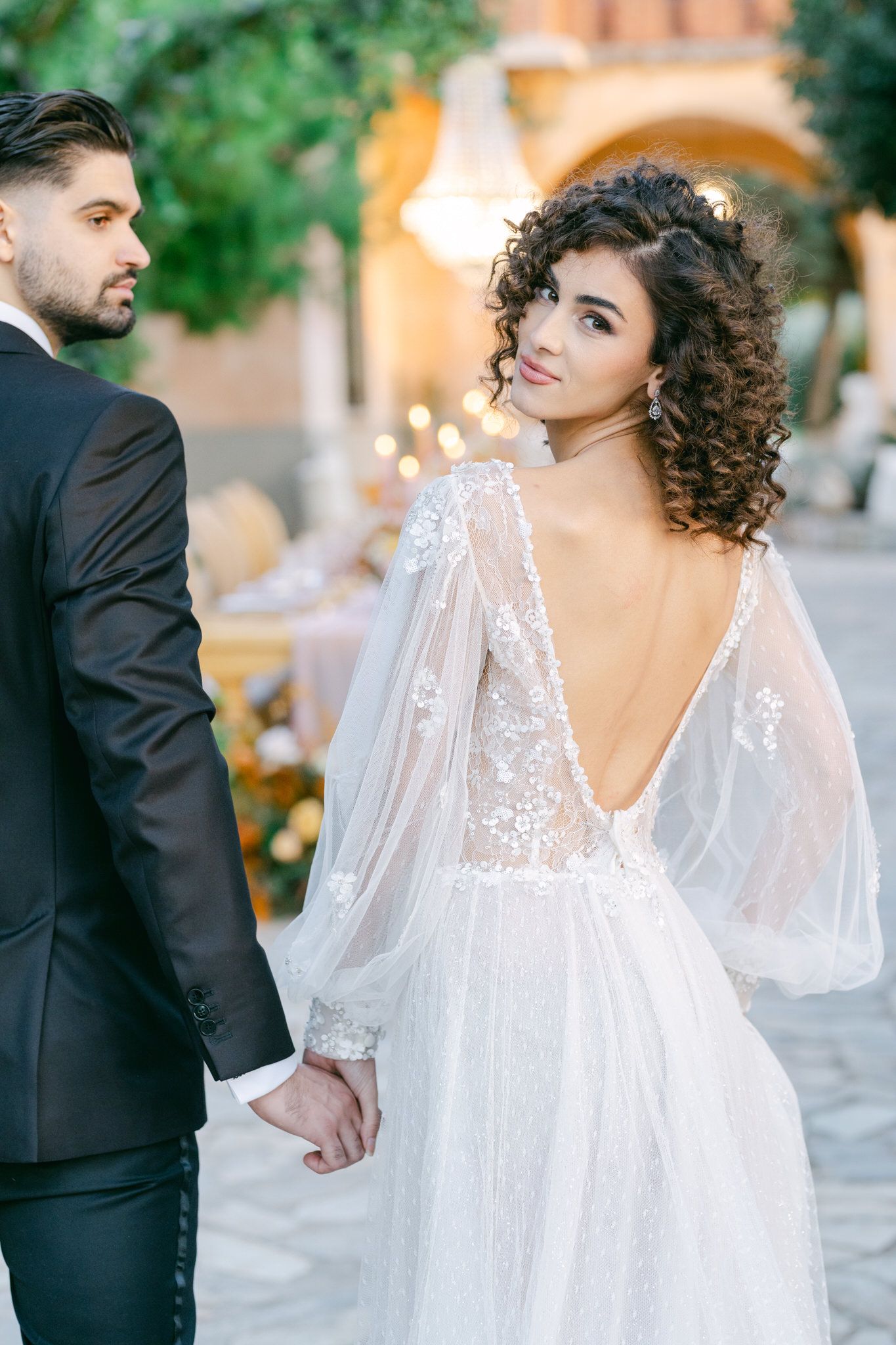 A romantic wedding photo showing a bride in a flowing white dress with sheer sleeves and open back with a groom in black suit.