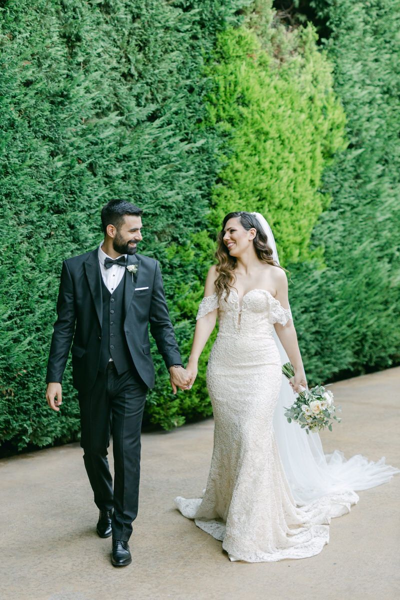 A bride in a lace wedding gown and groom in a black tuxedo walk hand in hand along a garden path with green hedges.