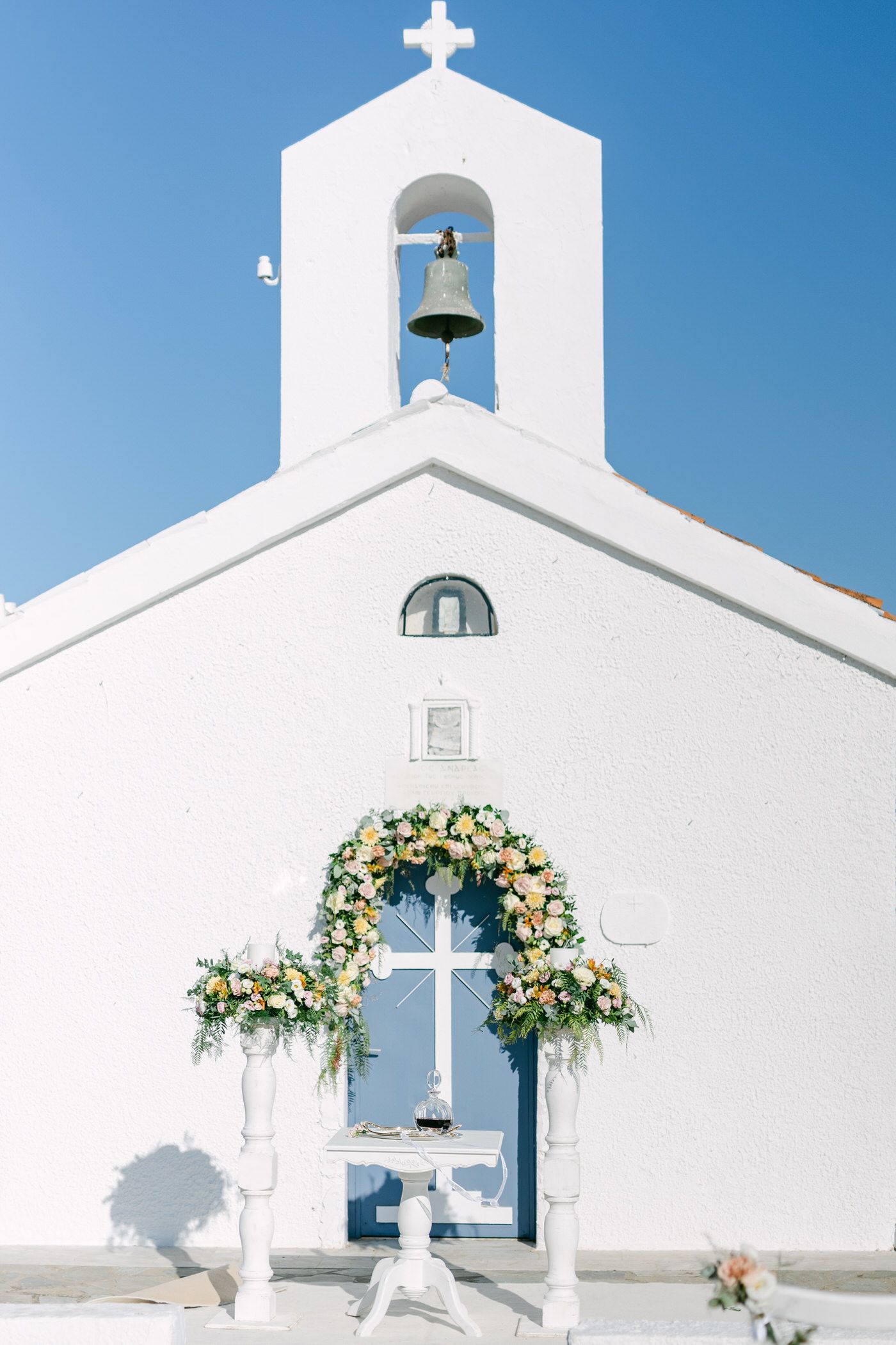 Simple white Greek church with bell tower decorated with floral arch and cross at entrance against bright blue sky.