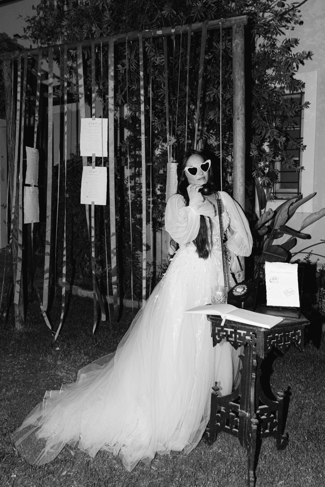 A figure in a flowing white wedding dress and sunglasses poses outdoors next to a decorative table in black and white. Κτήμα Λιάκος