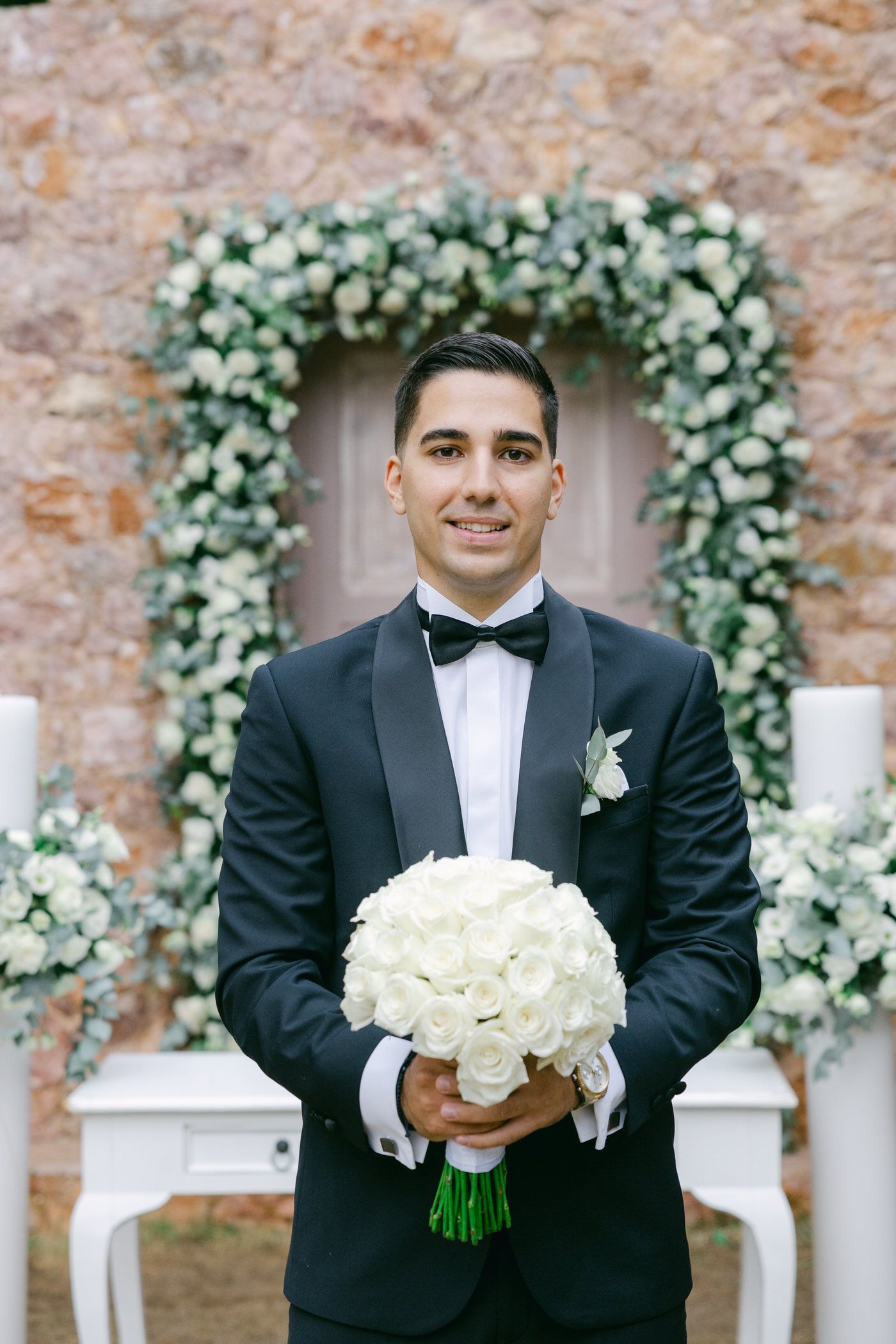 Classic wedding scene with floral arch and formal wear against rustic stone wall backdrop.