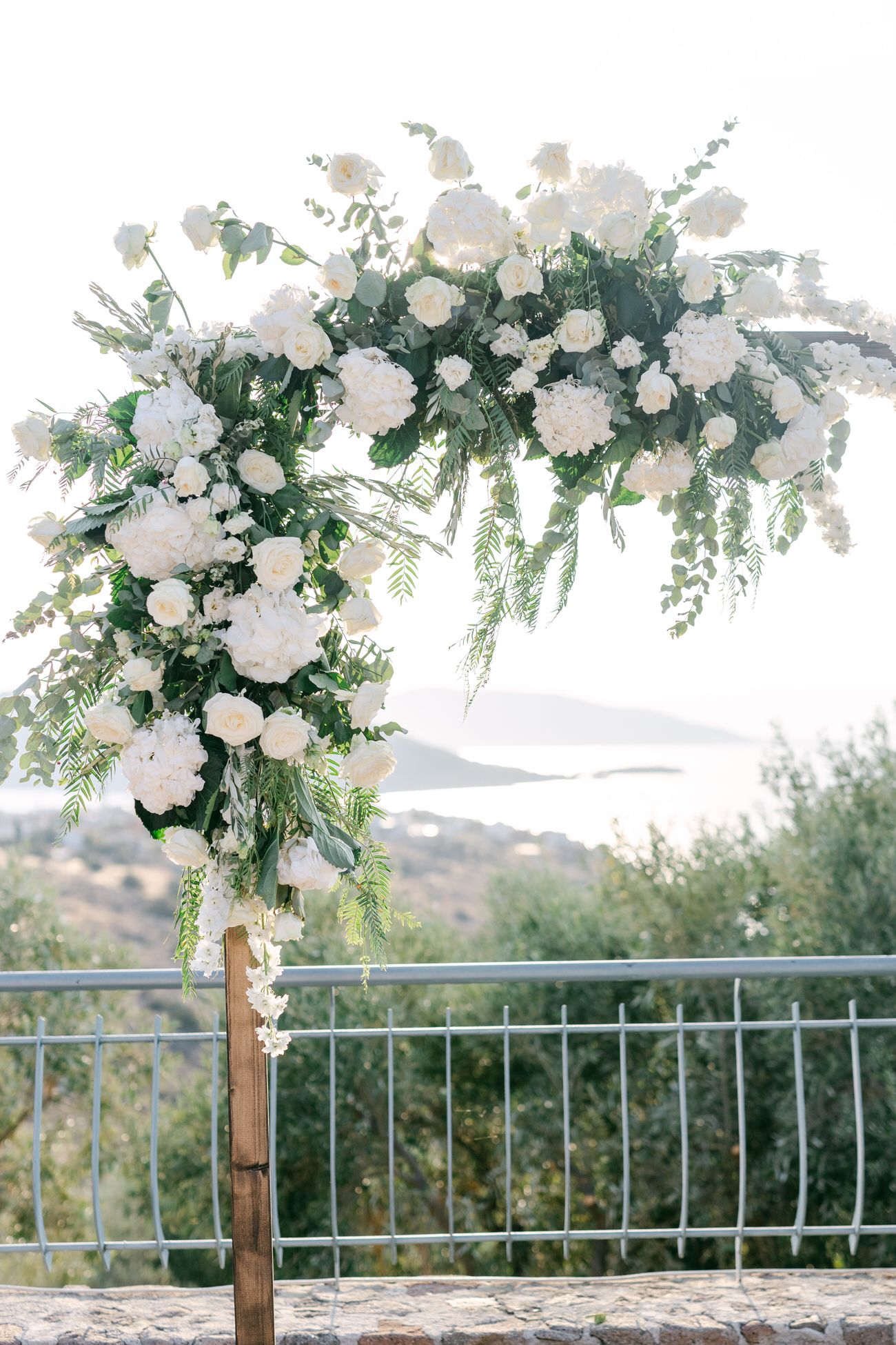 Elegant white floral wedding arch with cascading flowers and greenery against mountain landscape view.
