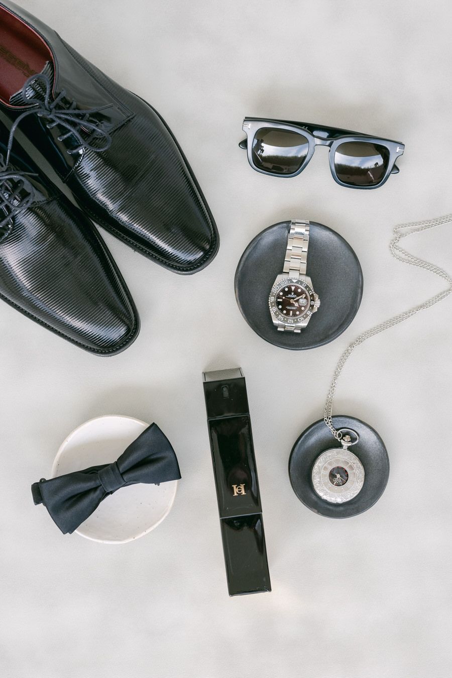 Elegant groom's accessories including black dress shoes, sunglasses, watch, bow tie, and leather belt arranged on white surface.