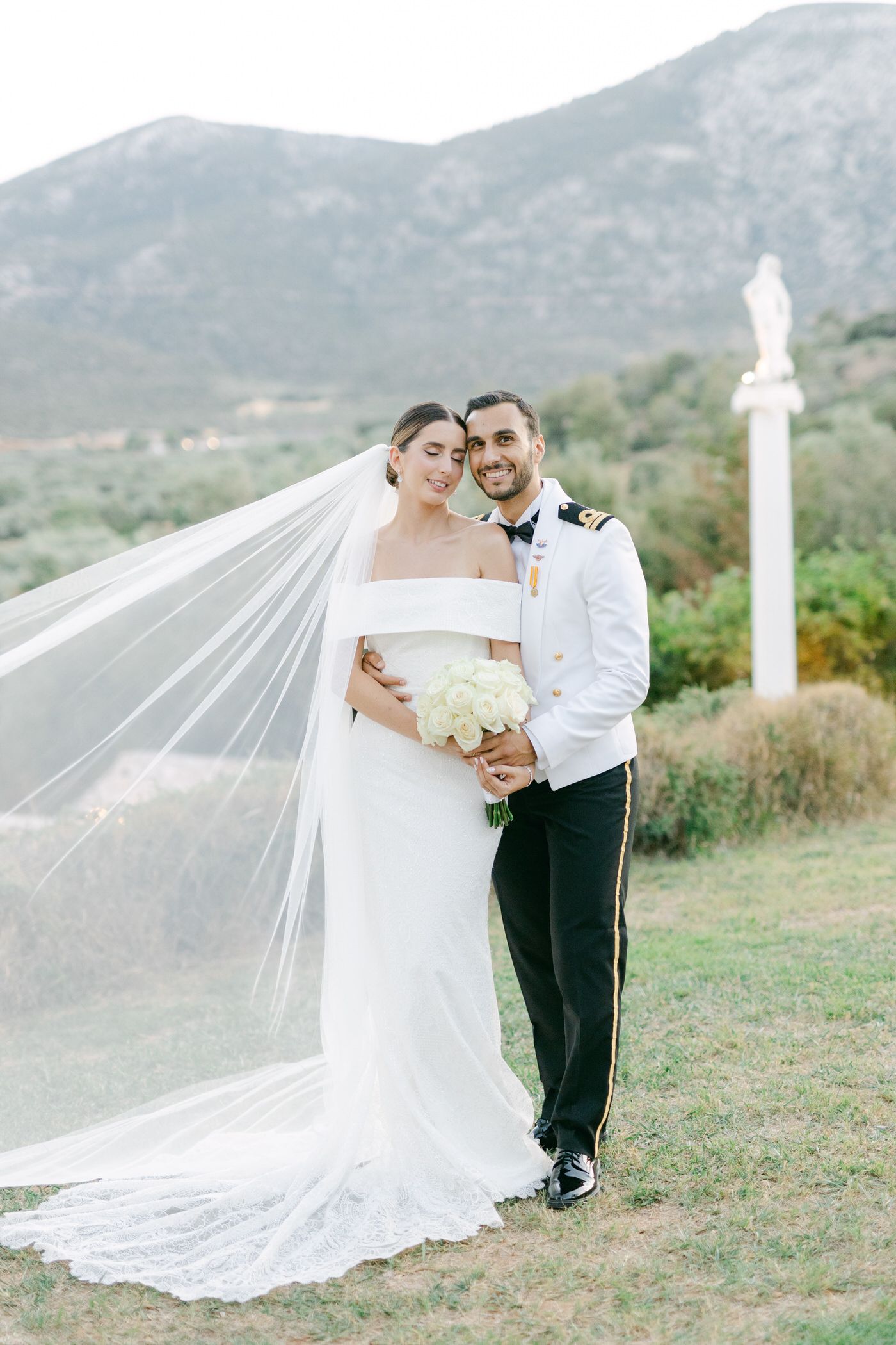 A couple in wedding attire poses outdoors with mountains in the background and a flowing white veil catching the breeze.