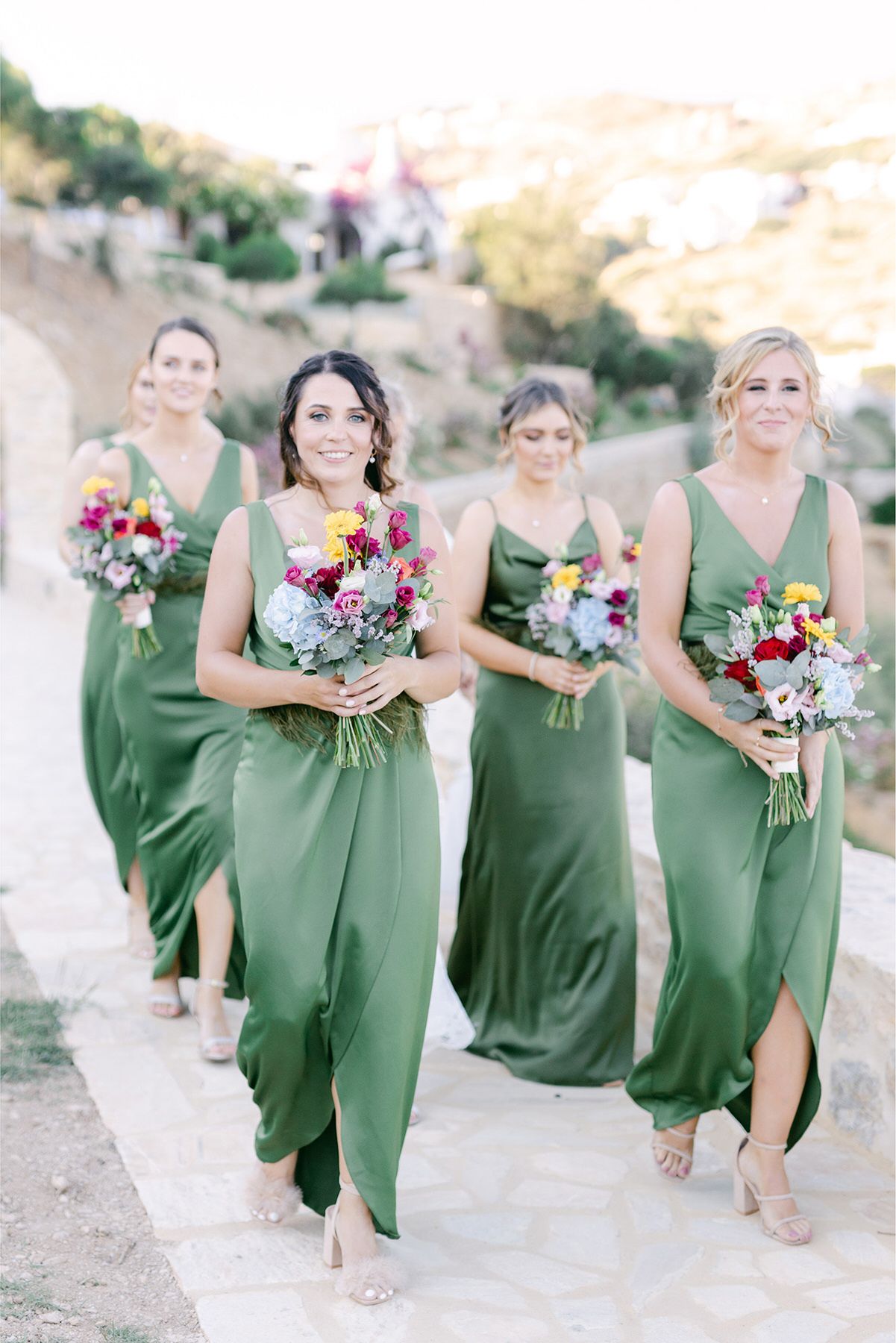 Bridesmaids in matching sage green dresses walk outdoors carrying vibrant floral bouquets at a wedding.
