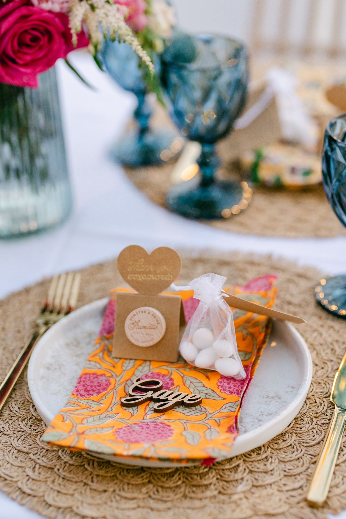 Wedding place setting with orange floral napkin, heart-shaped favor box and blue glassware on gold charger plate.