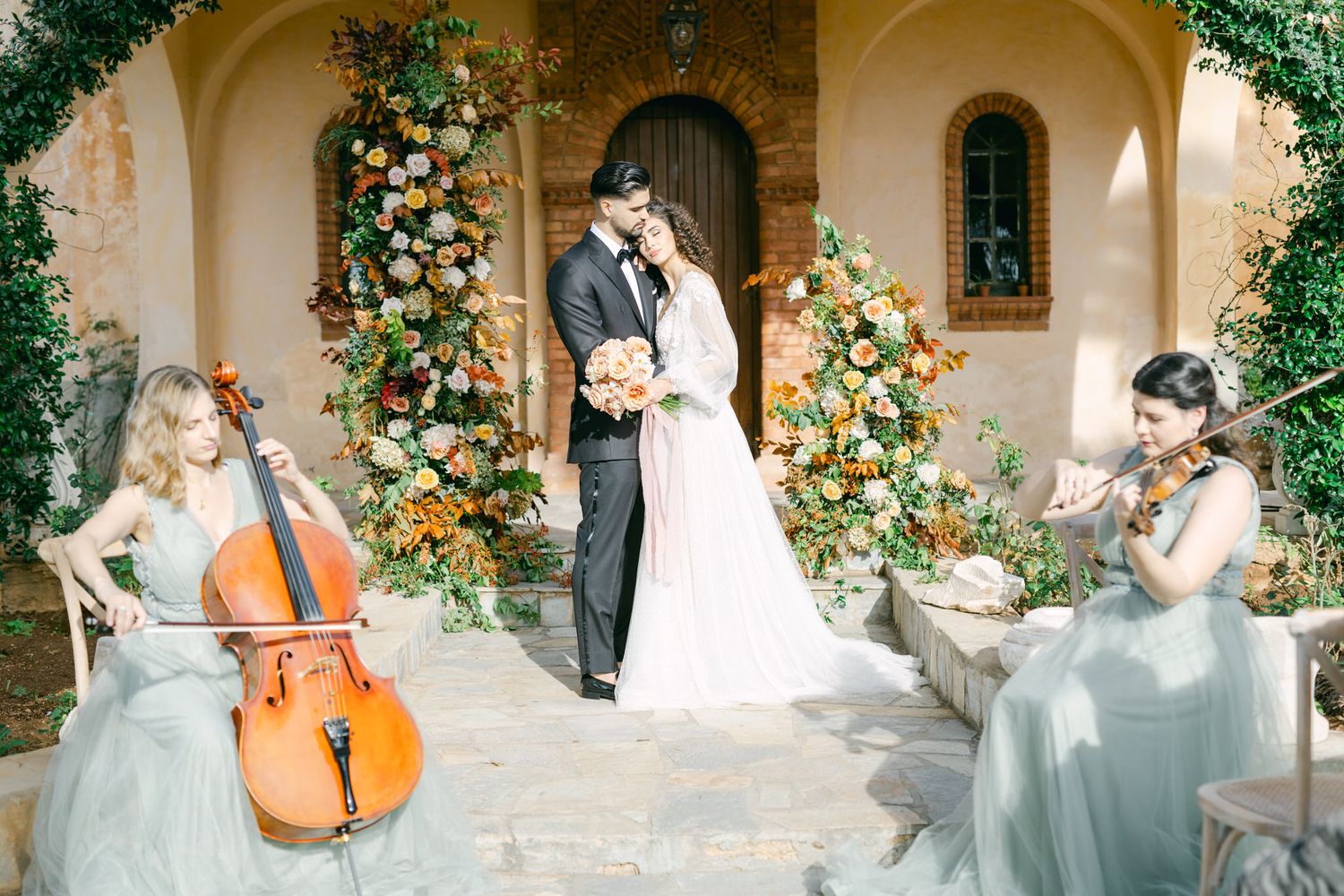 Elegant outdoor wedding scene with musicians playing cello and flute amidst floral decorations at a villa entrance.