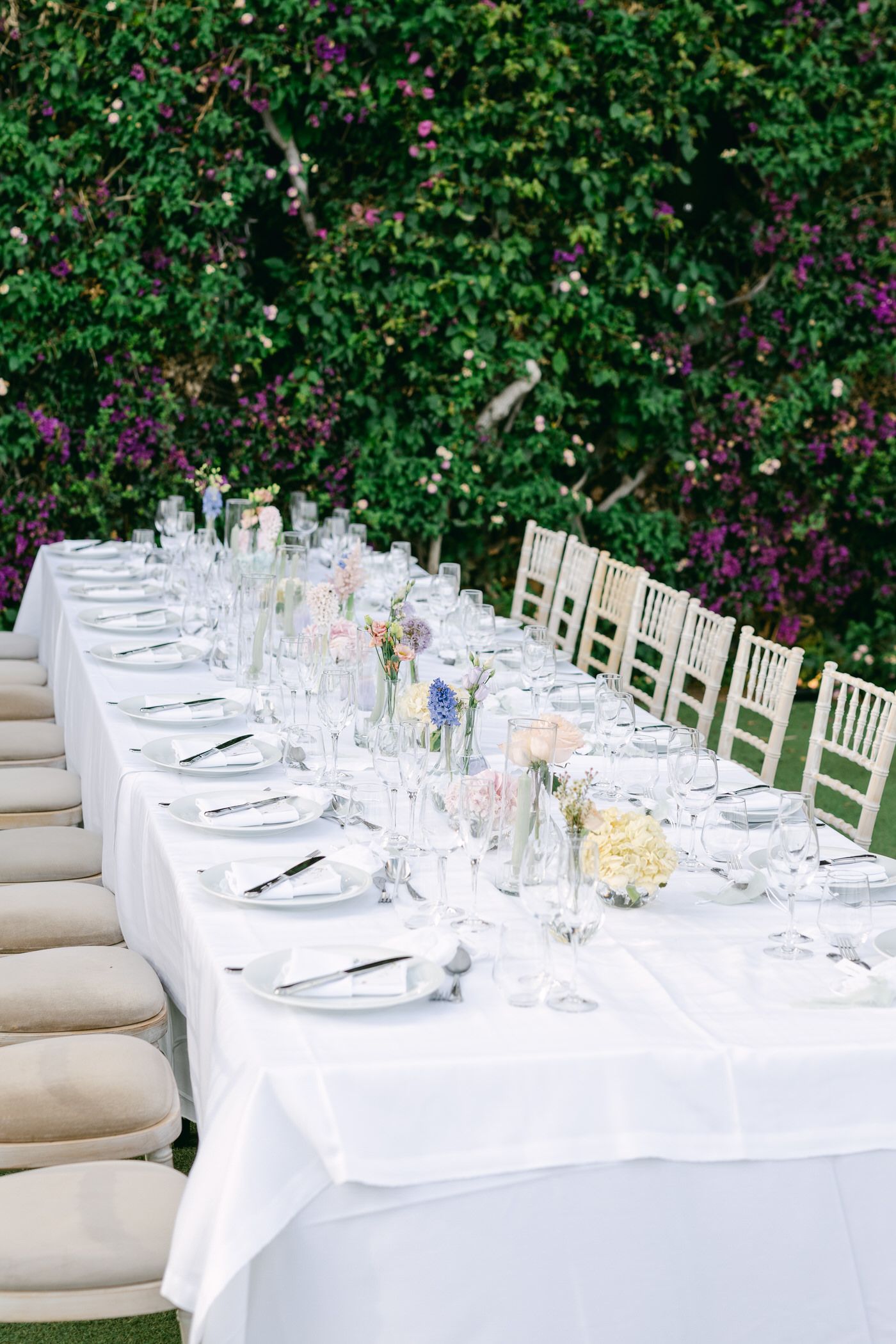 Elegant outdoor wedding table setting with white linens, Chiavari chairs and floral centerpieces against lush garden backdrop.