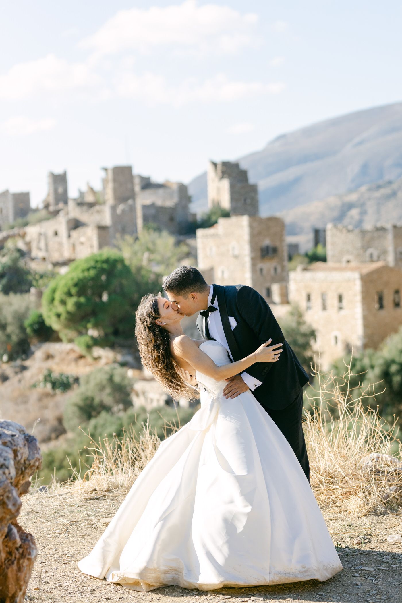Wedding couple shares romantic moment in front of ancient stone buildings and mountains in Mediterranean setting.