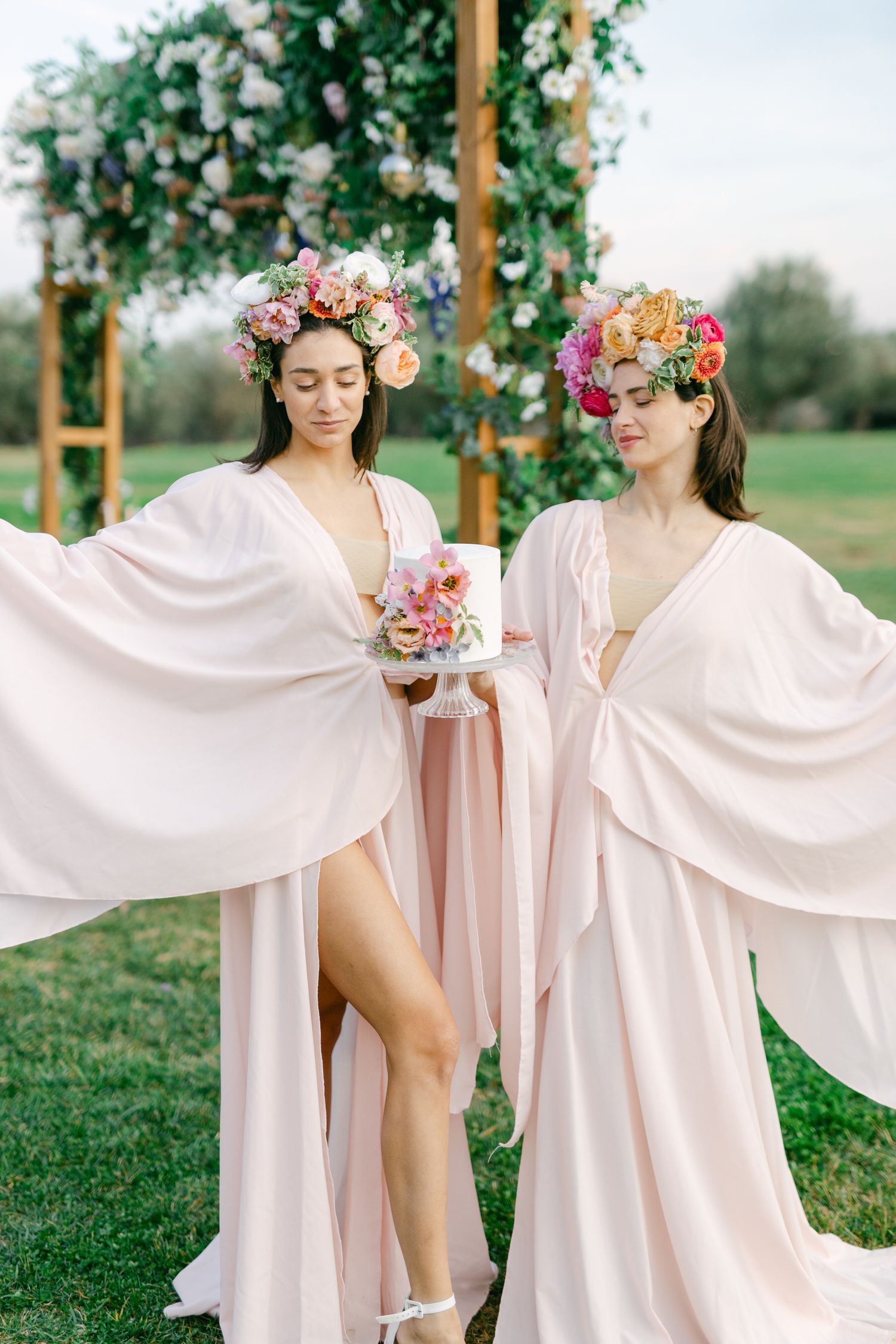 Ethereal garden scene with flowing blush pink robes and floral crowns amidst greenery and wooden posts.