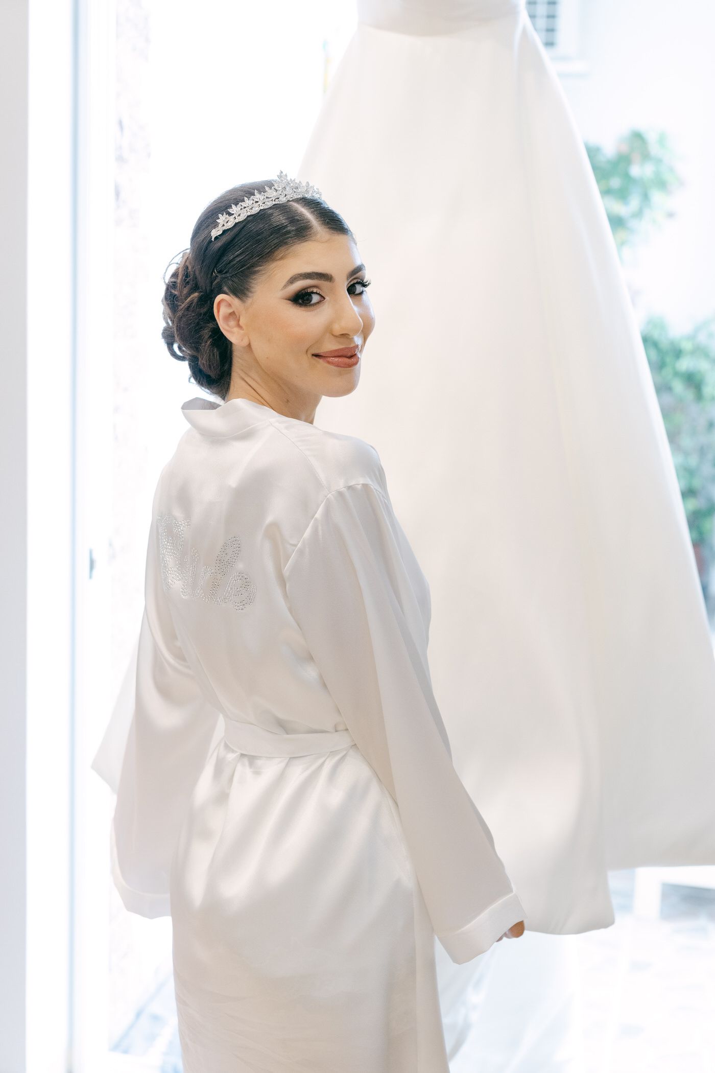 Woman in a white silk robe smiles elegantly on her wedding day morning wearing a delicate tiara.