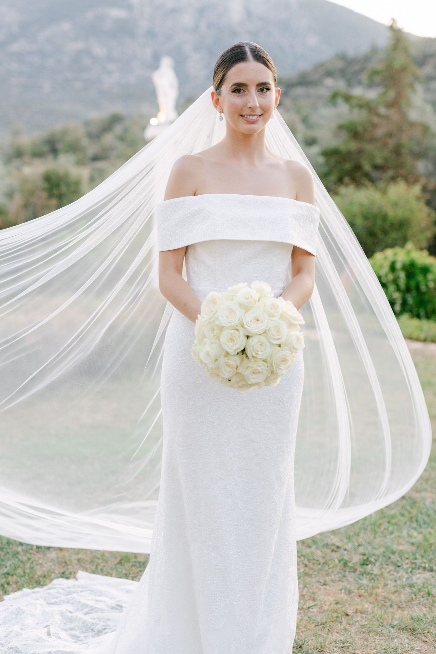 Elegant off-shoulder wedding dress with long flowing veil and white rose bouquet against mountain backdrop.