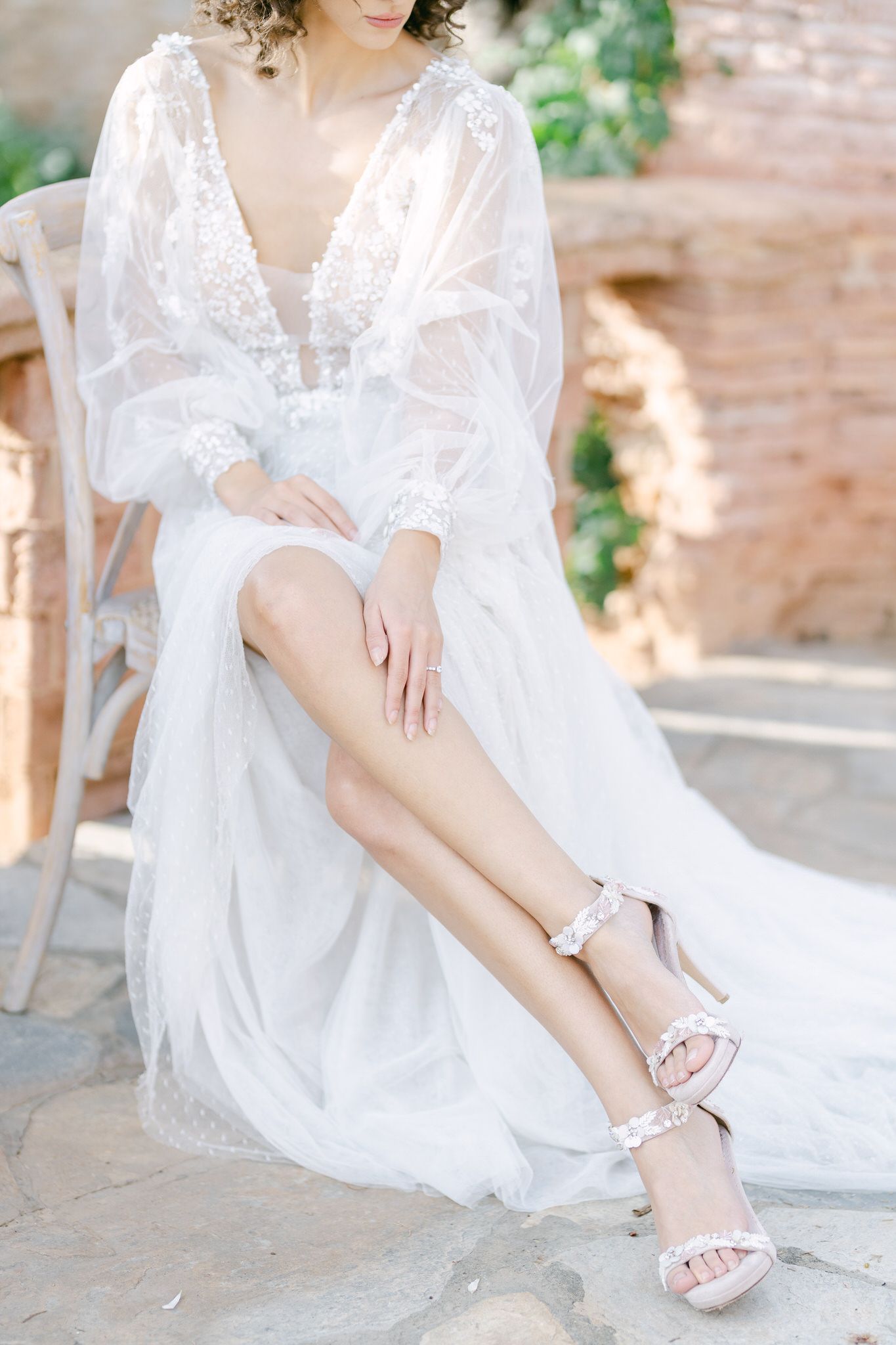 Delicate white lace wedding gown with flowing sleeves and elegant sandals shown in a romantic bridal portrait.