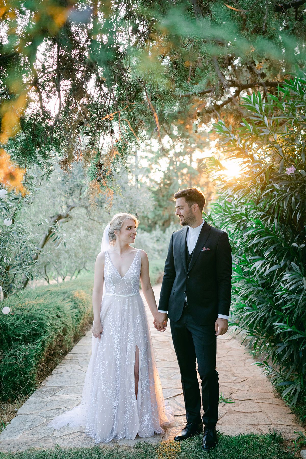 A couple walks hand in hand along a stone path surrounded by lush greenery and dreamy sunlit trees.
