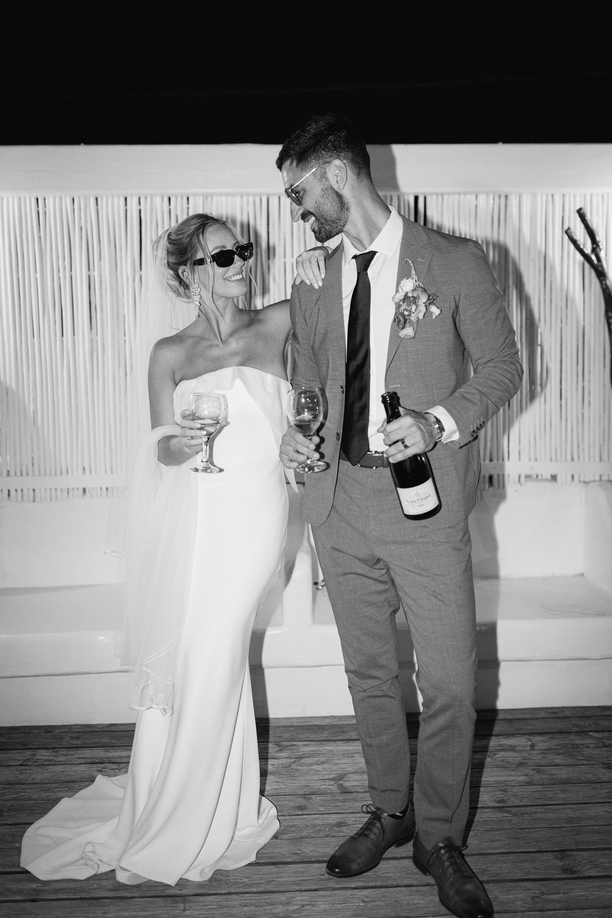 Black and white photo of a couple sharing champagne at their wedding reception, bride in white dress and groom in grey suit.