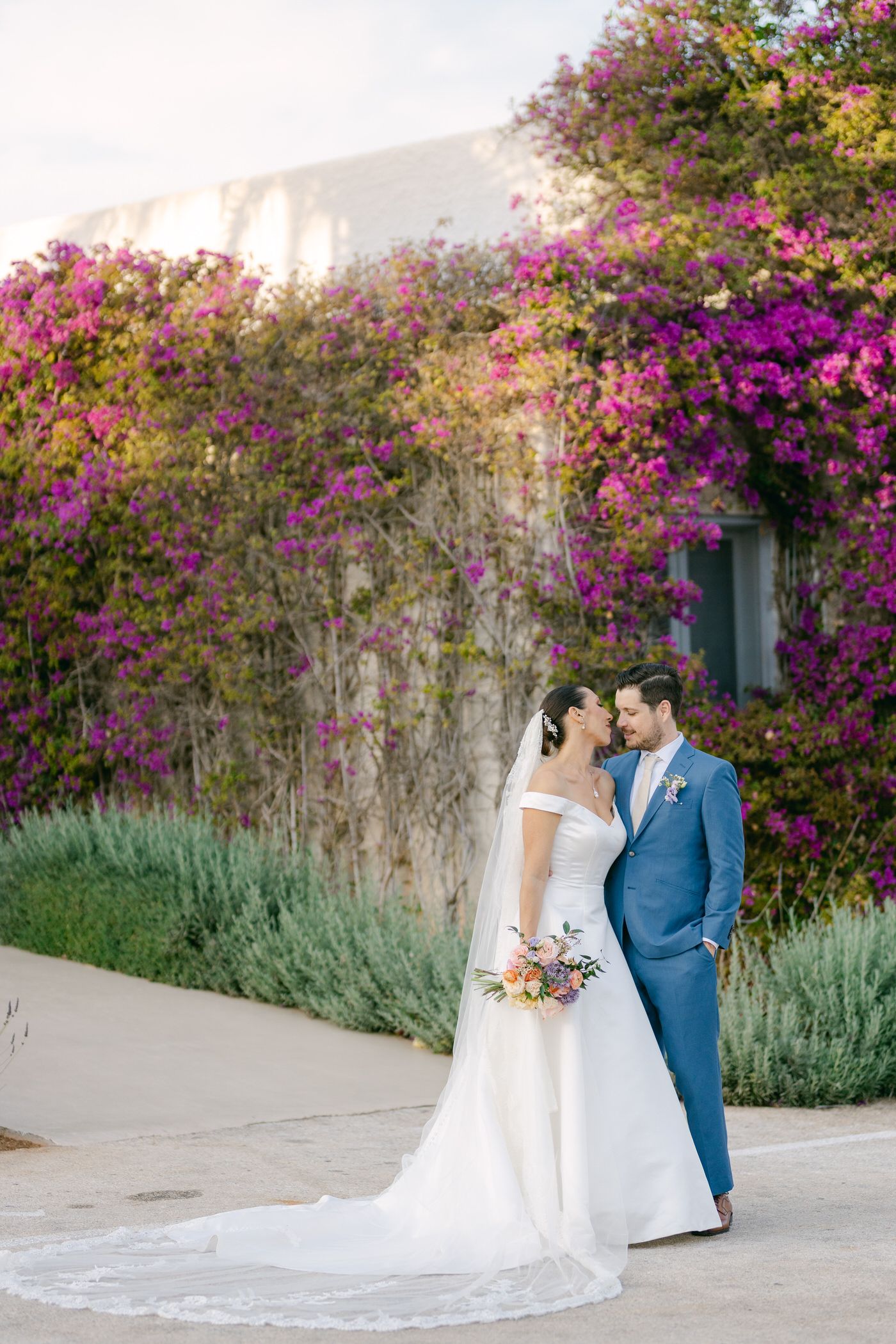 A couple stands together against a wall covered in vibrant pink bougainvillea flowers and green foliage.