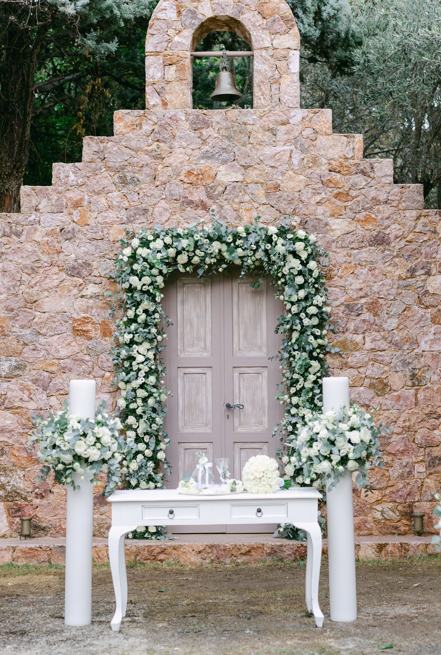 Elegant rustic stone chapel entrance adorned with white floral arch and vintage table with candles for a romantic wedding setup.