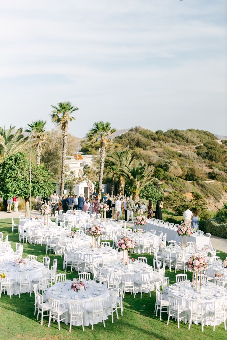 Round reception tables with white chairs set up on a lawn with palm trees in the background.