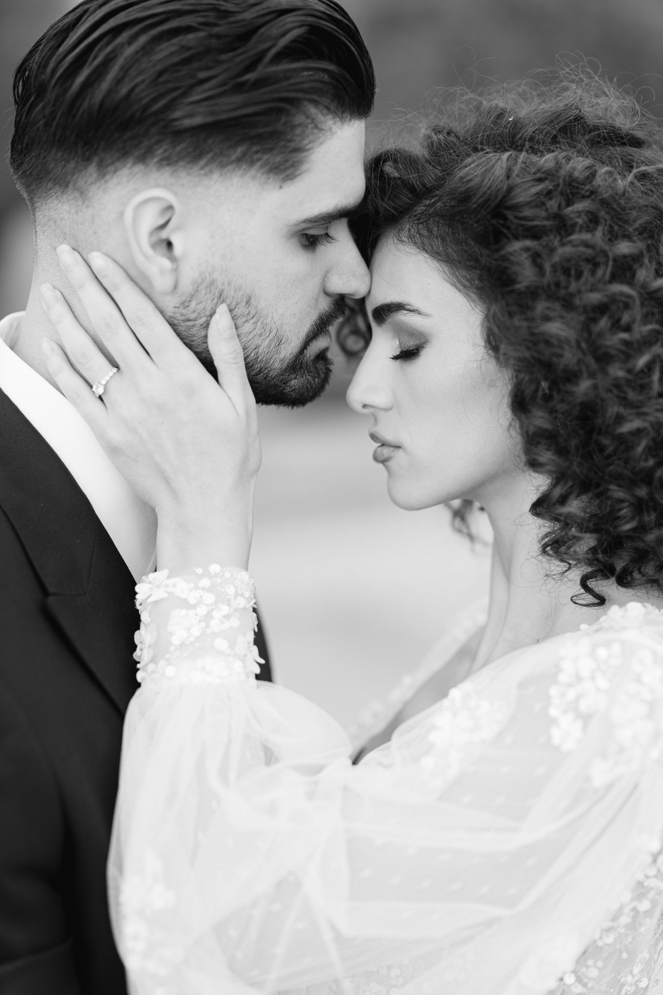 Romantic black and white wedding portrait showing tender moment between couple, bride in lace gown with curly hair.