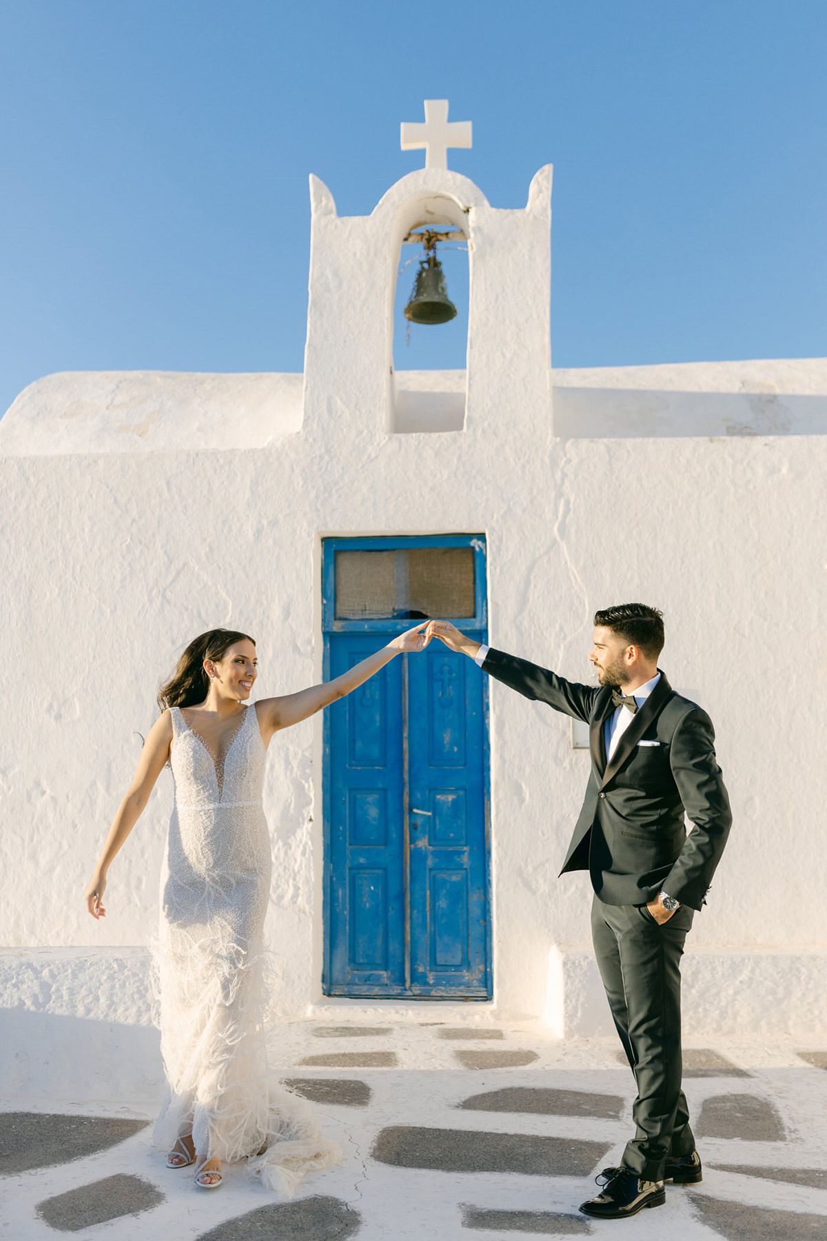A couple in wedding attire dancing outside a traditional white Greek church with a blue door and bell tower in Santorini.