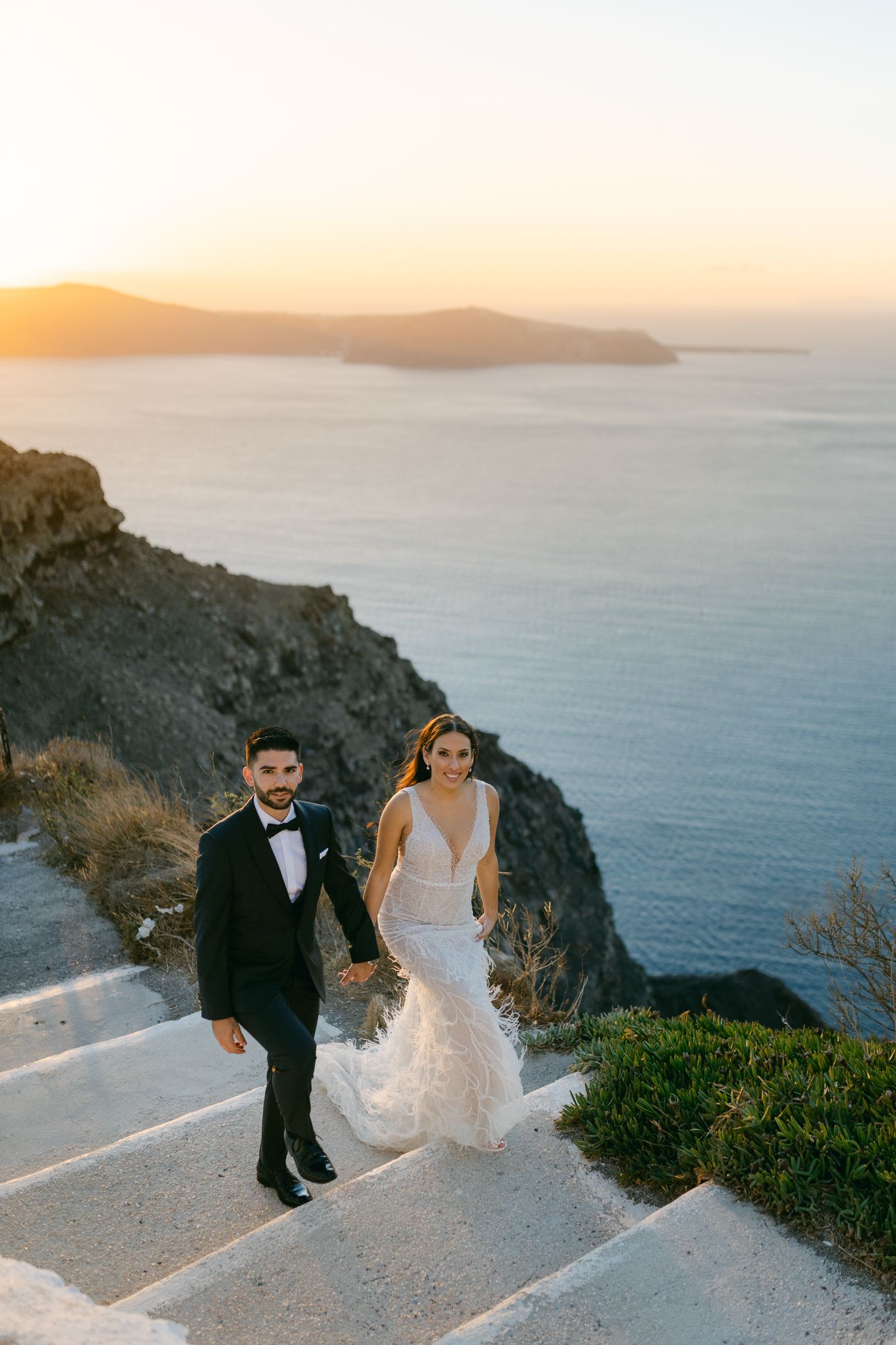 A couple poses for wedding photos against a stunning sunset view of the Mediterranean Sea from cliffs in Santorini, Greece.