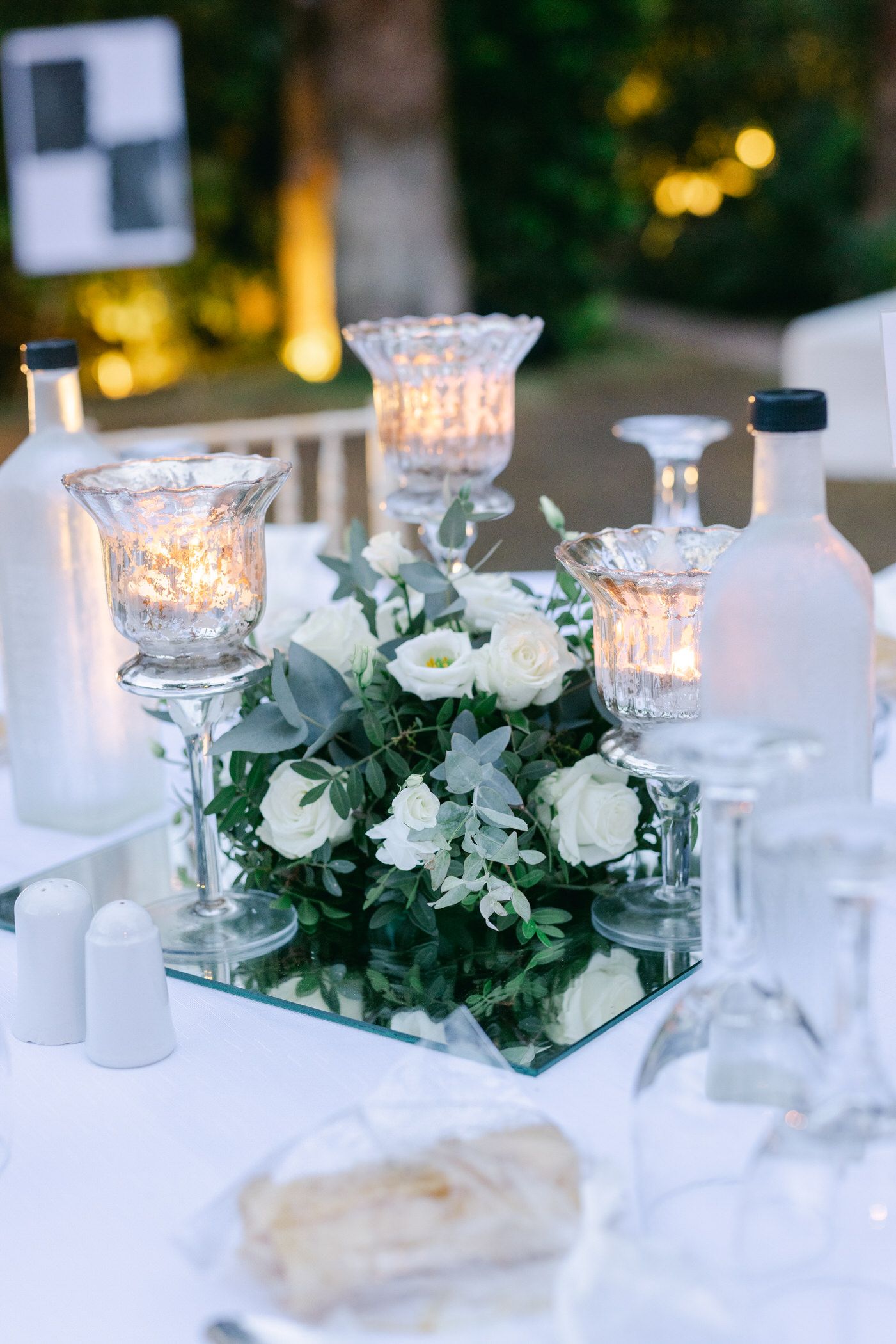 Elegant wedding table centerpiece with white roses and candlelit mercury glass holders on a mirrored base.