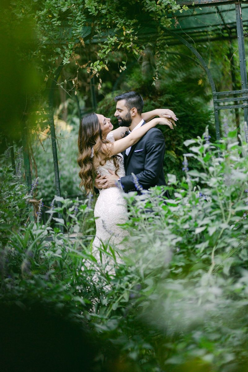 A romantic couple embraces in a lush garden setting with greenery and natural light filtering through the foliage.