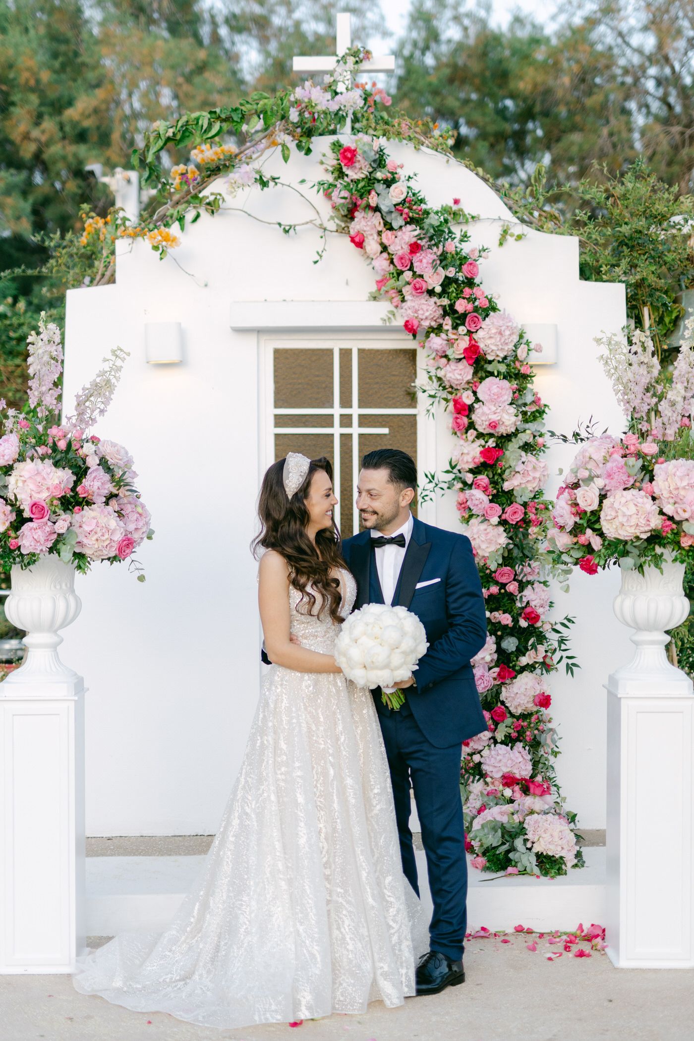 Wedding couple under white arch decorated with pink roses and greenery.